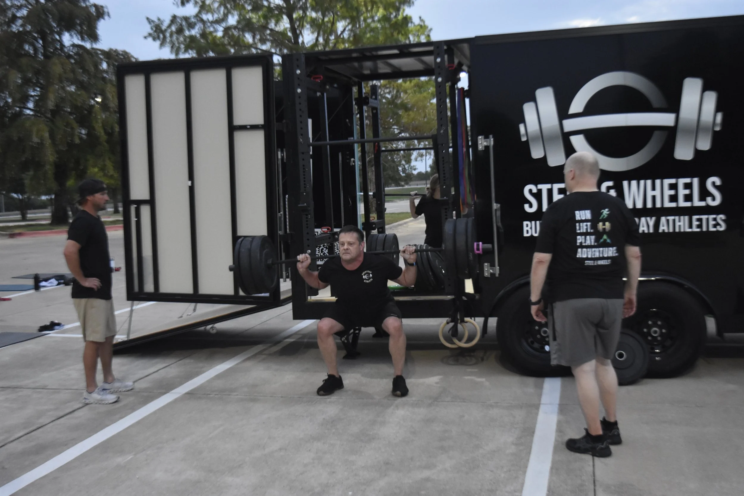 A man is performing a squat exercise with a barbell on his shoulders, while other men observe near a mobile gym trailer with a large logo and the words "Steel Wheels" and "Burpee. Run. Lift. Play. Adventure." written on it, outdoors in a parking lot.
