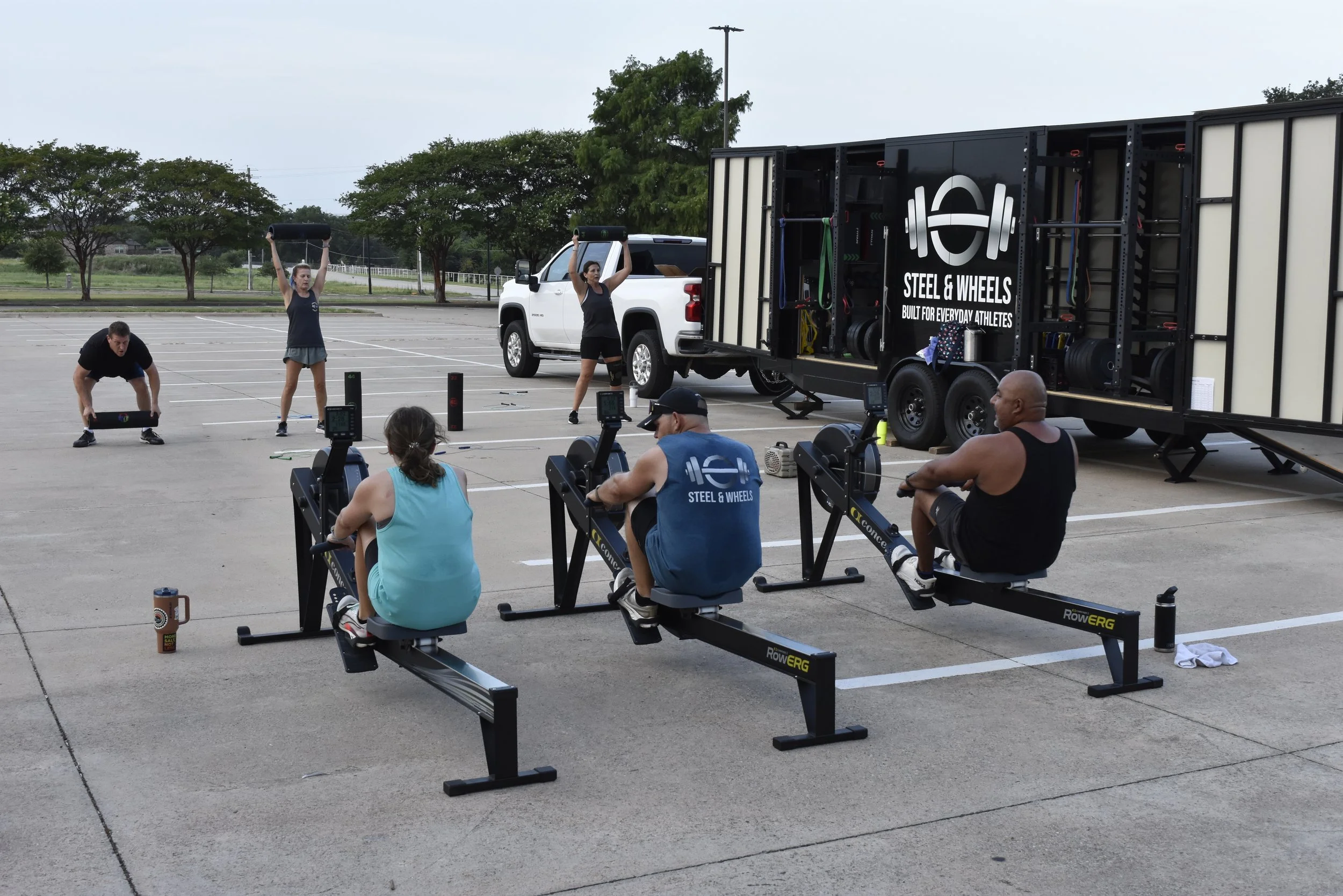 Group of people participating in a workout session outdoors, with some using rowing machines and others doing exercises, set up in a parking lot near a large mobile fitness trailer.