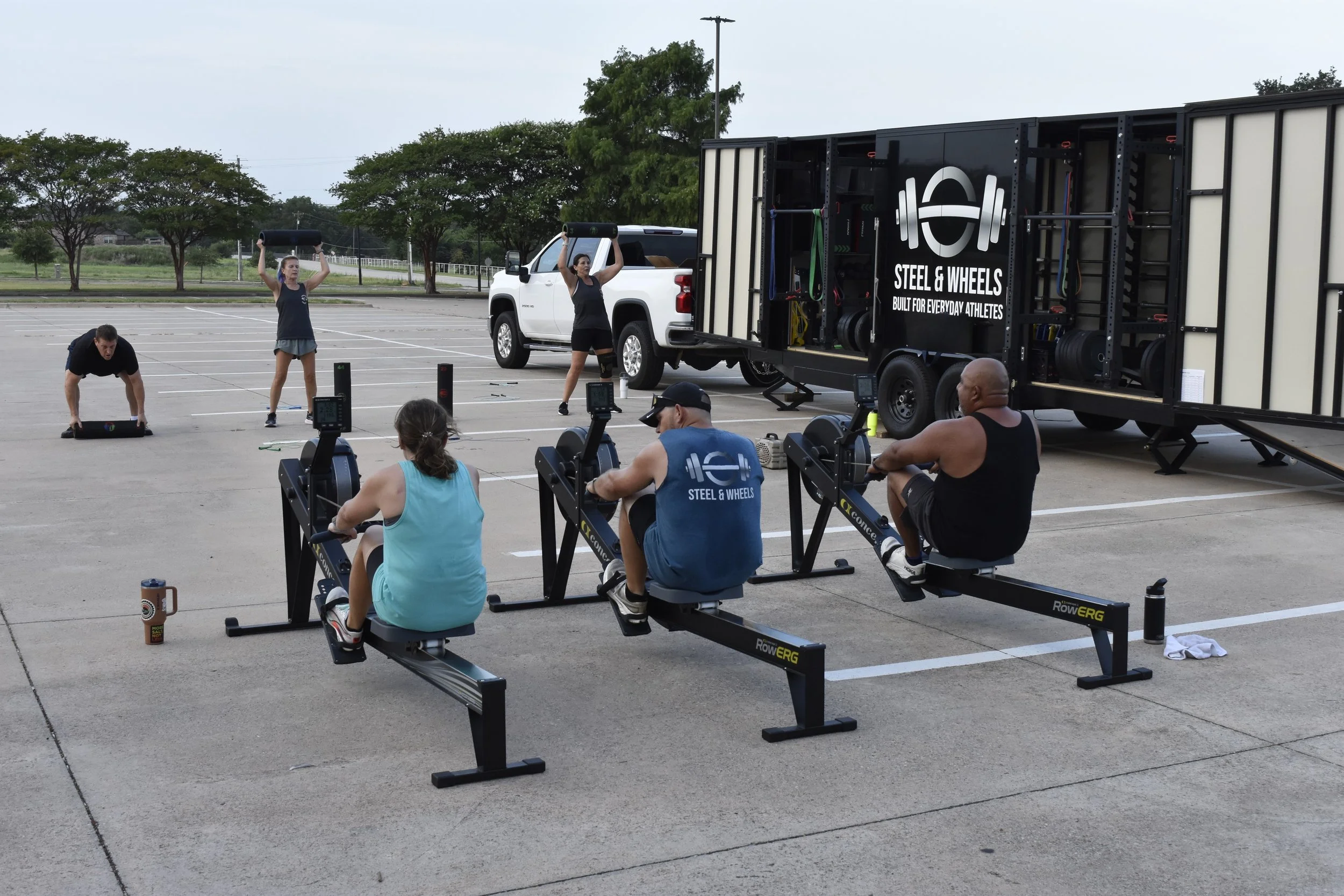 People participating in an outdoor fitness class in a parking lot, using exercise equipment and weights with a truck labeled 'Steel & Wheels' in the background.