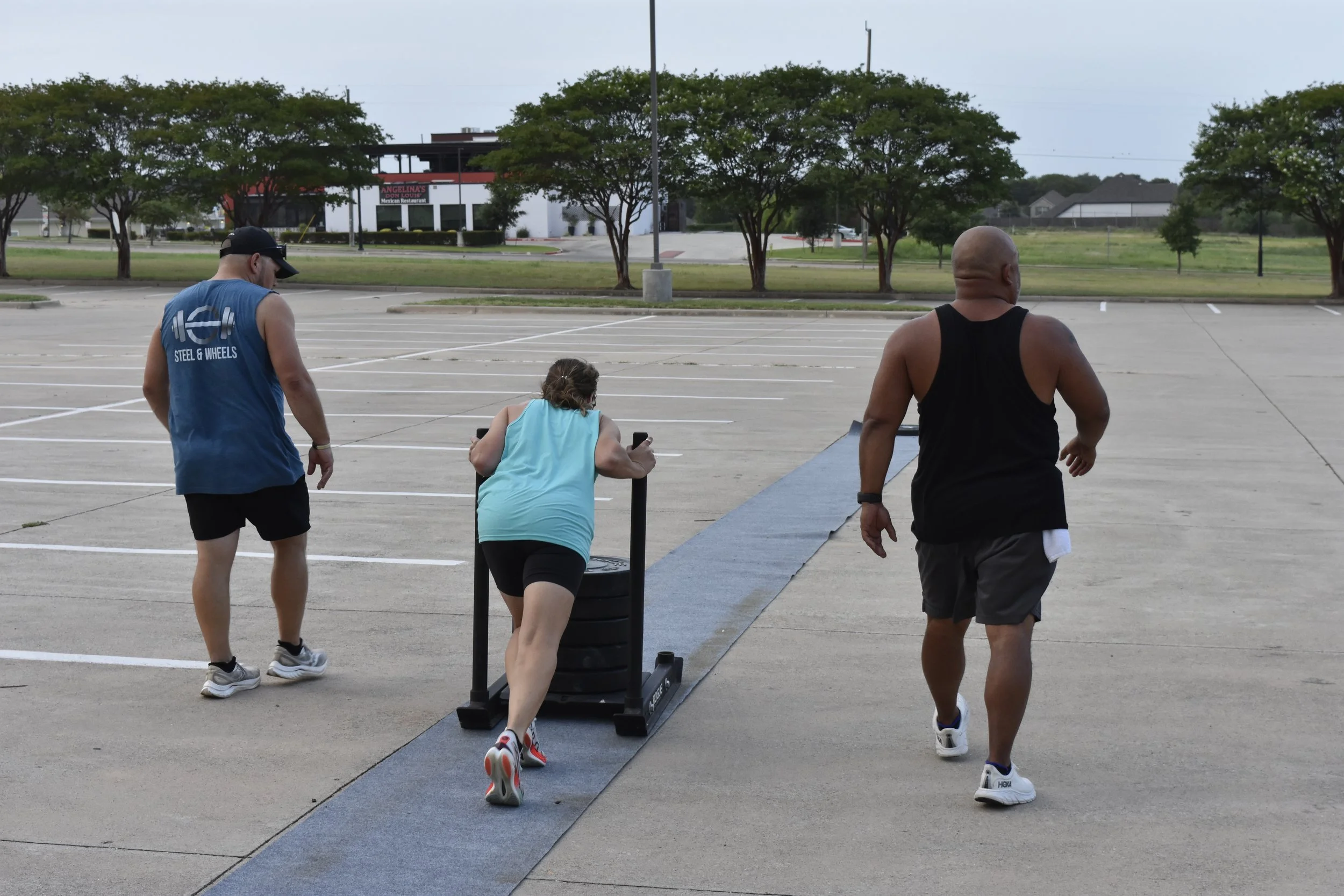 Three people working out outdoors in a parking lot, with trees and a restaurant in the background. One woman is using a sled for exercise, while two men are running behind her.