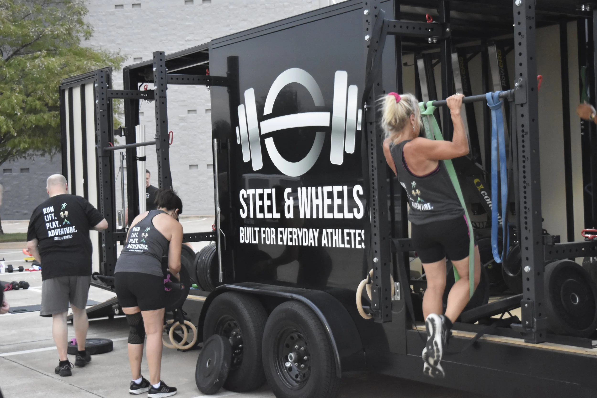 Women exercising on a mobile gym unit with weights and resistance bands outside. The side of the unit has text that reads, 'Steel & Wheels. Built for everyday athletes.'