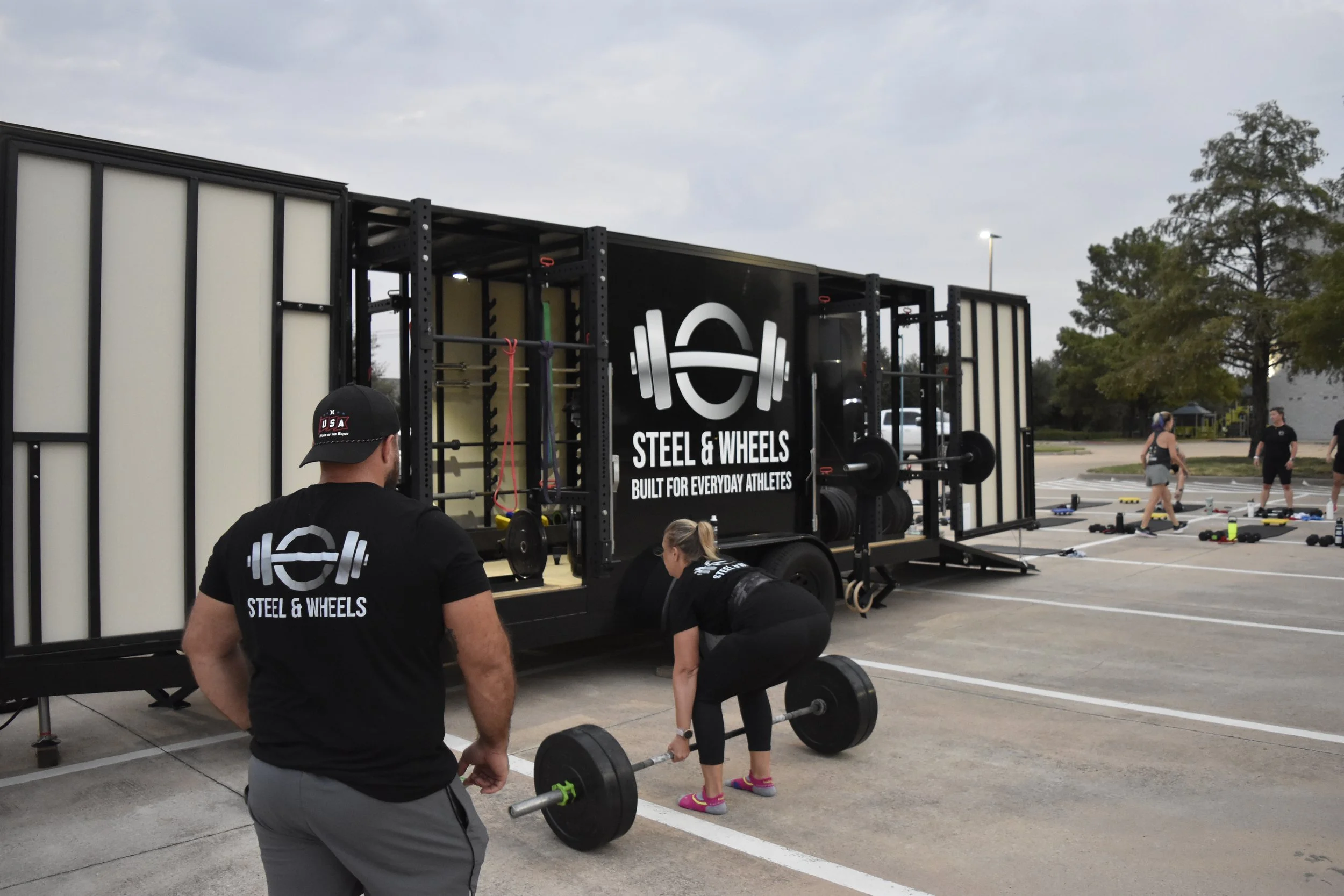 People exercising outside in a parking lot during a fitness event, with a mobile gym setup and various workout equipment.