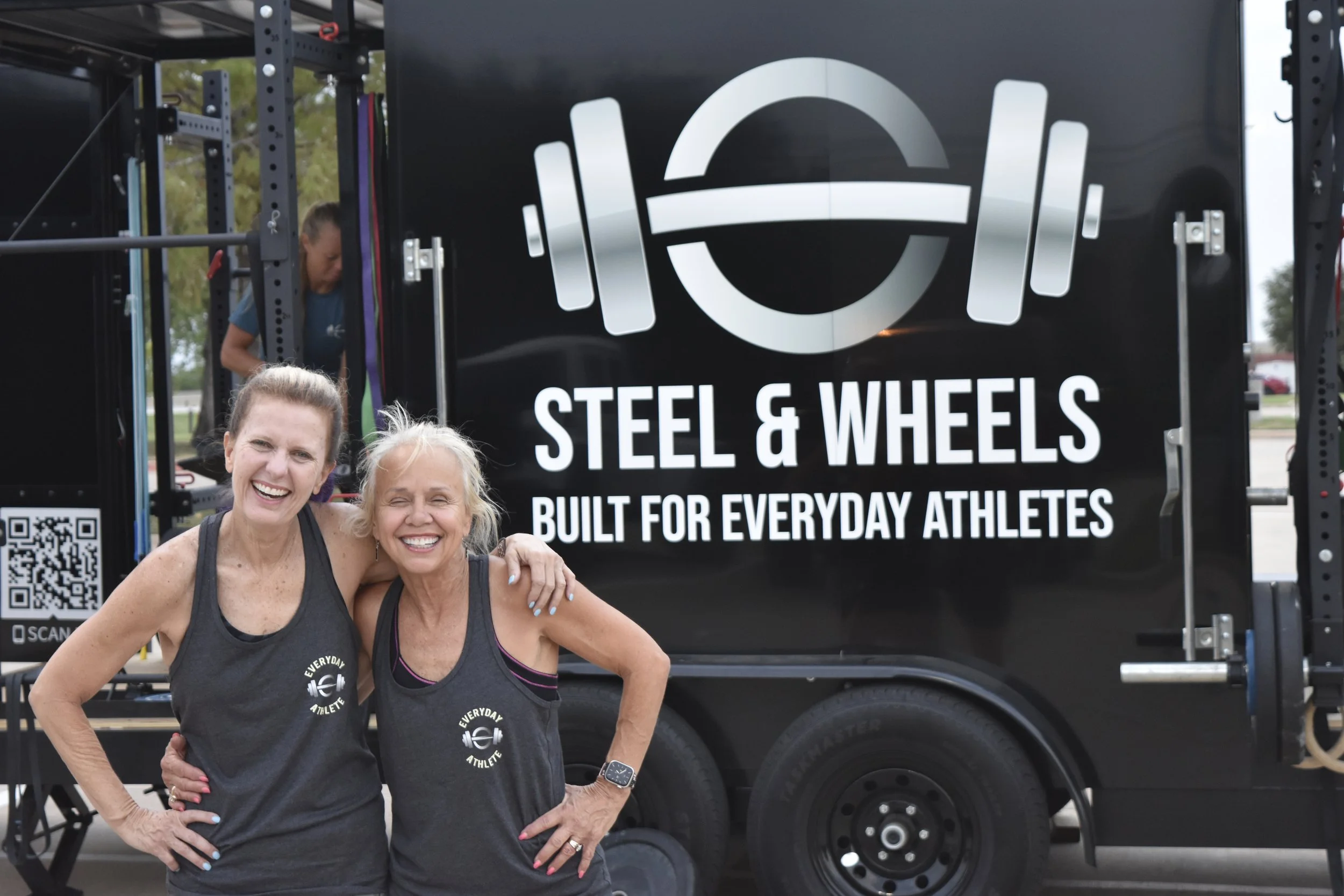 Two women smiling and standing together in front of a gym truck with 'Steel & Wheels' logo, wearing black workout shirts that say 'Everyday Athletes'.