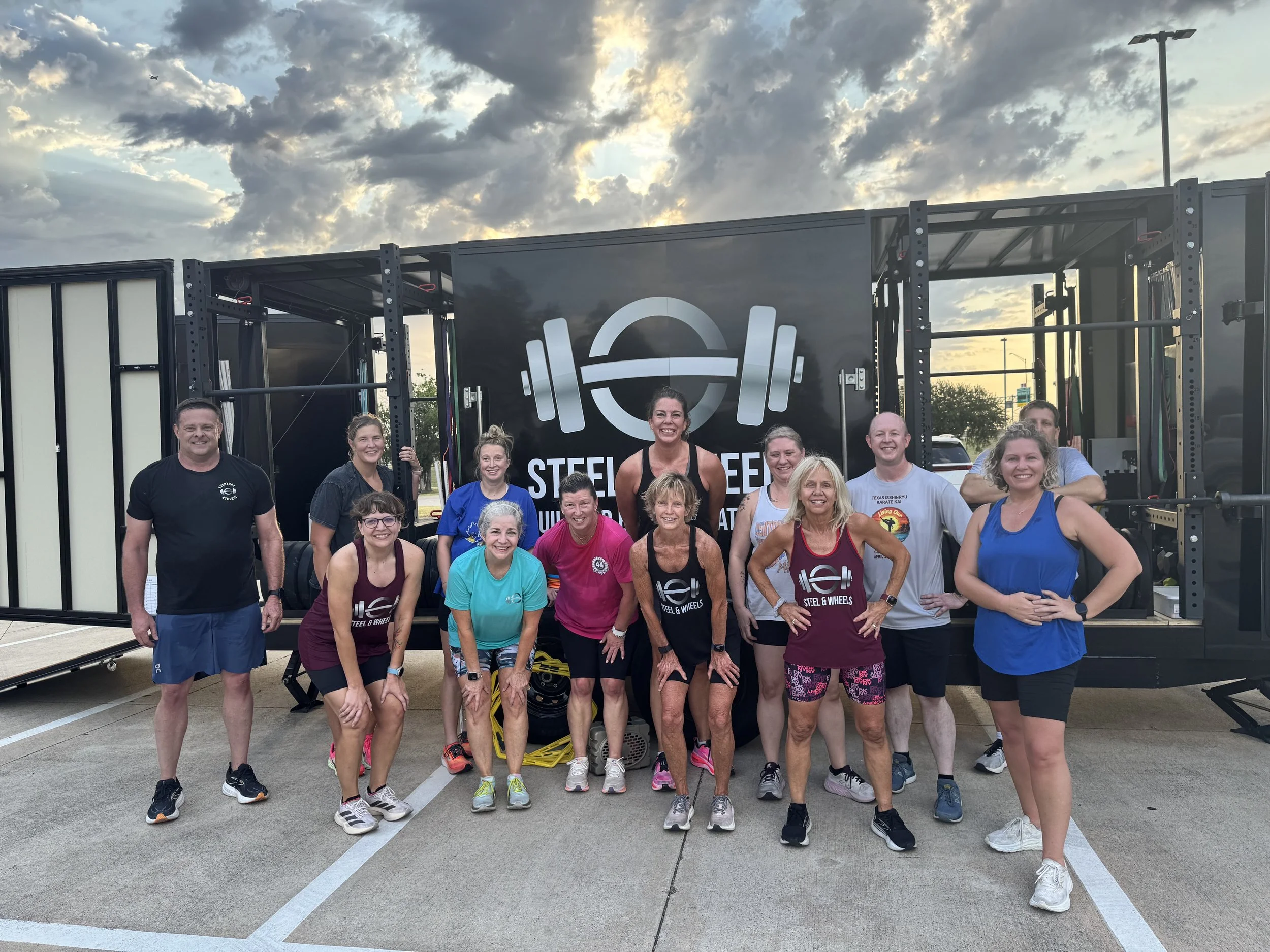 Group of people in athletic clothing posing in front of fitness equipment outdoors on a cloudy day.