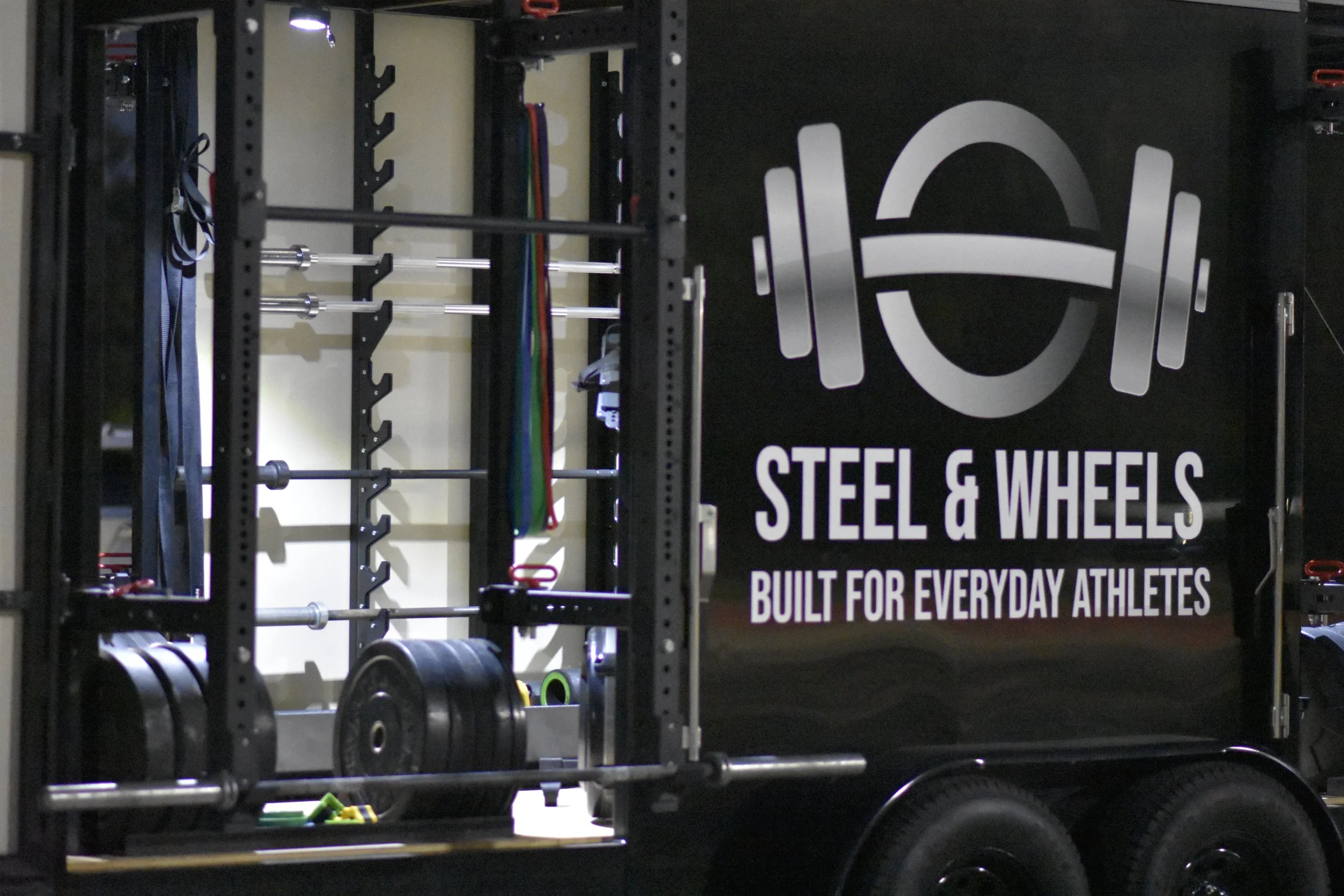 A black mobile gym equipment cart with a large logo and the words "Steel & Wheels" and "Built for everyday athletes." The cart contains various workout equipment like weight plates, barbells, and resistance bands.