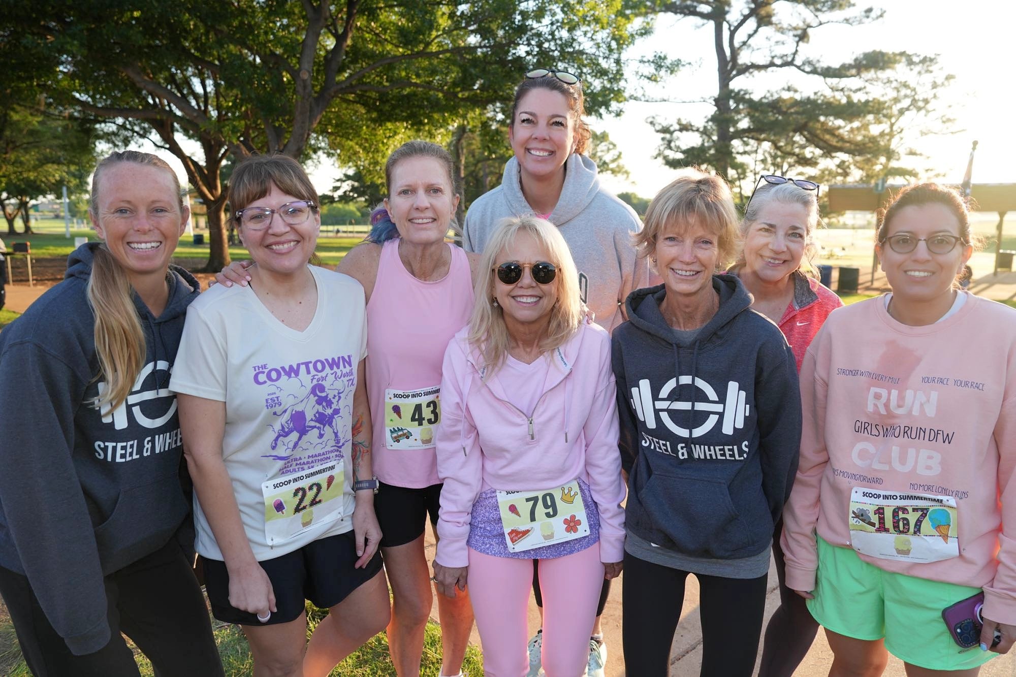 A group of ten women standing together outdoors in a park during a sunny day, smiling for the camera, with some wearing race bibs and athletic clothing.