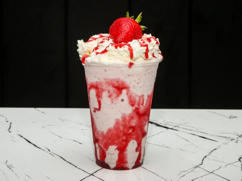Strawberry milkshake topped with whipped cream, strawberries, and strawberry syrup in a clear glass