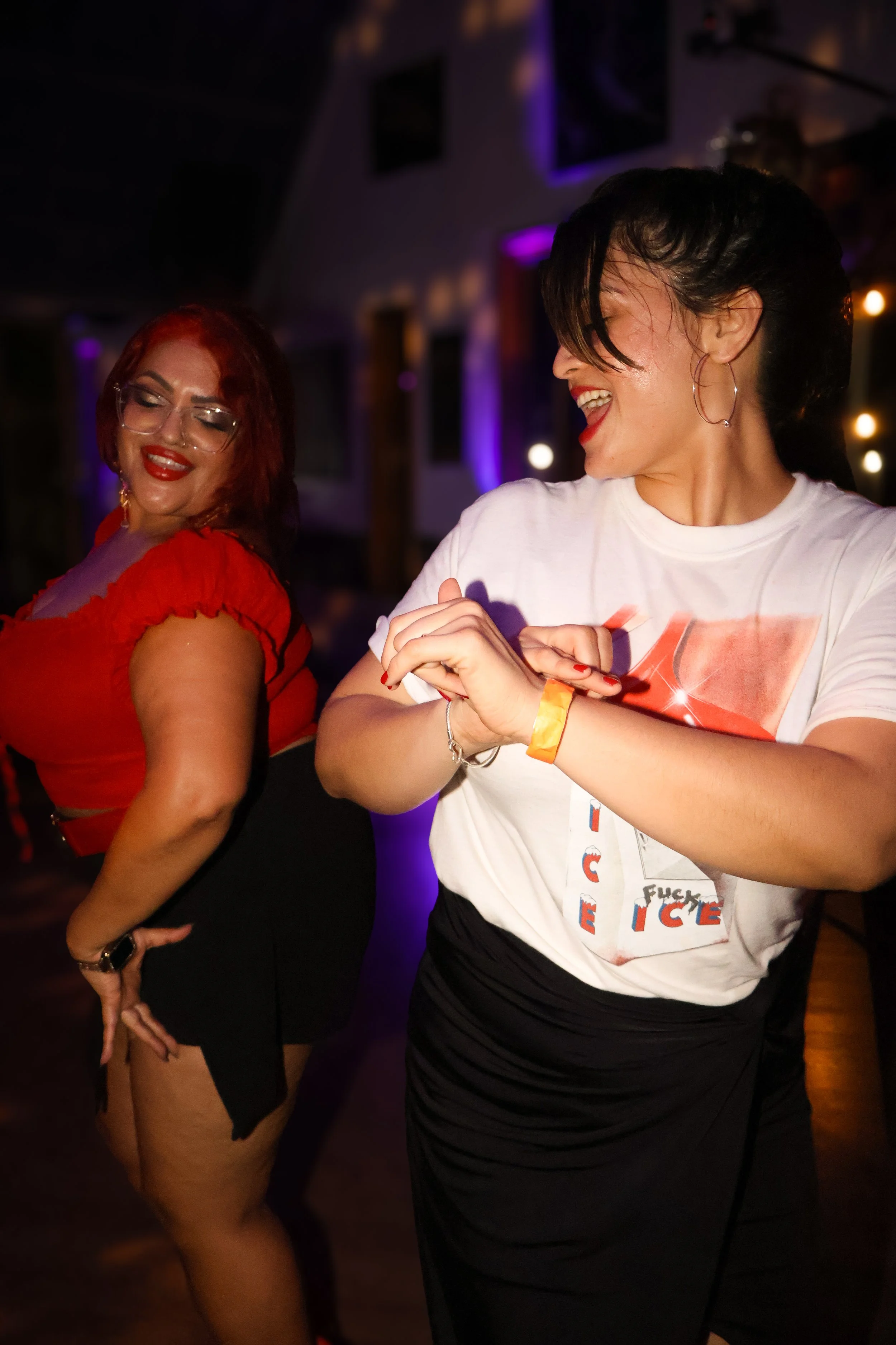 Dos mujeres bailando y riendo en un ambiente de fiesta con luces moradas y doradas.