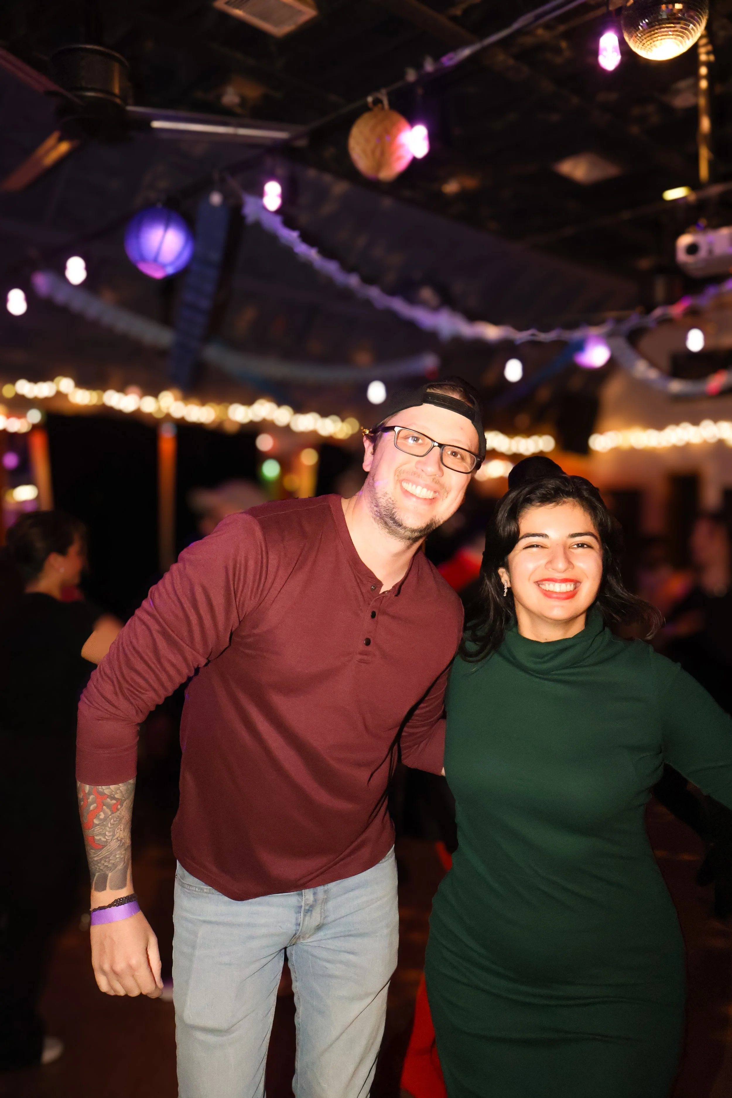 Dos personas sonriendo en un ambiente de fiesta, decoraciones con luces y adornos colgantes en el techo.