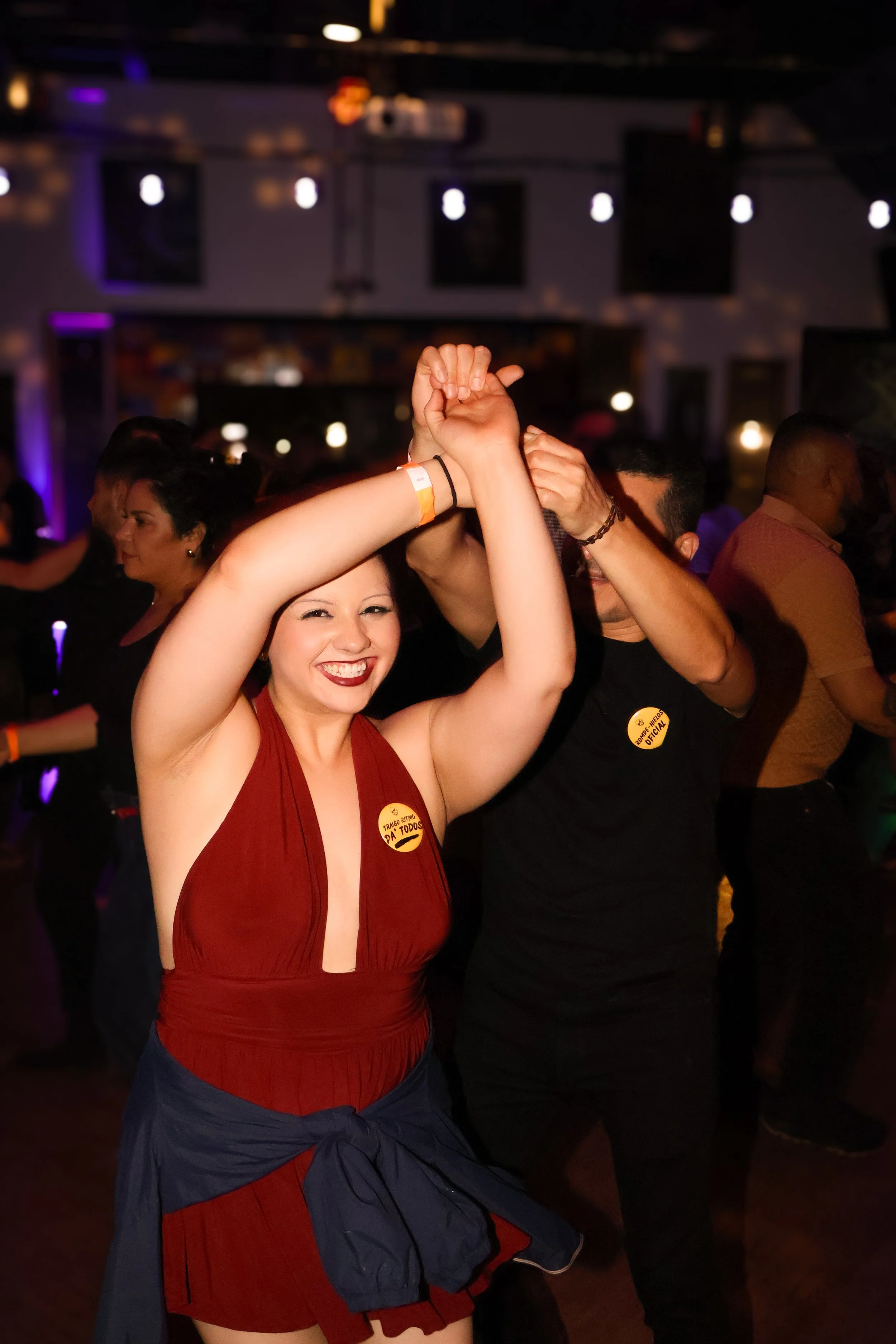 Joven mujer sonriendo mientras baila en una fiesta con fondo oscuro y otras personas bailando.