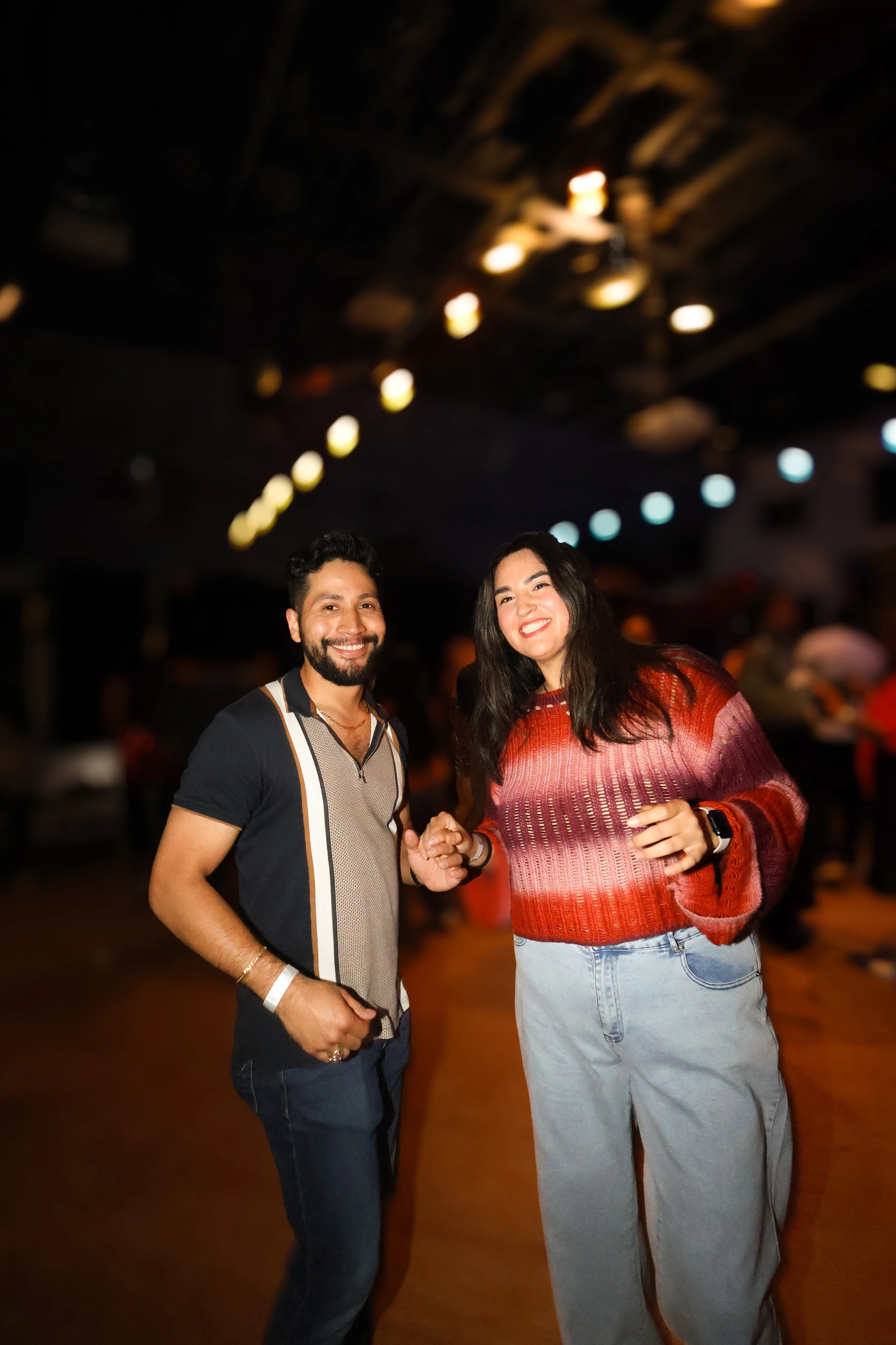Dos personas, un hombre y una mujer, sonriendo y dándose la mano en un lugar con poca iluminación y luces de techo difusas.