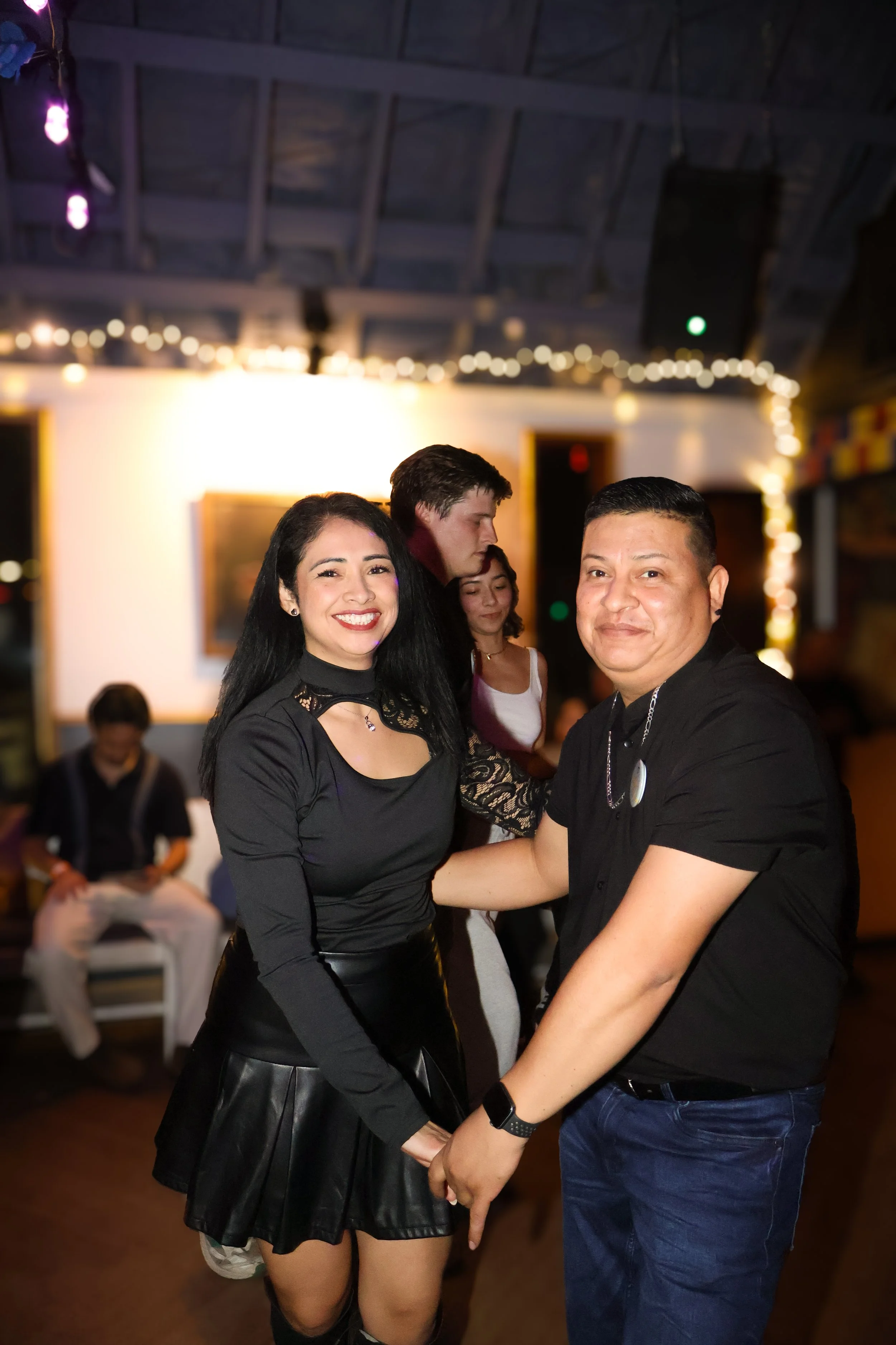 Una pareja bailando y sonriendo en un ambiente festivo, con personas en el fondo y decoraciones de luces.