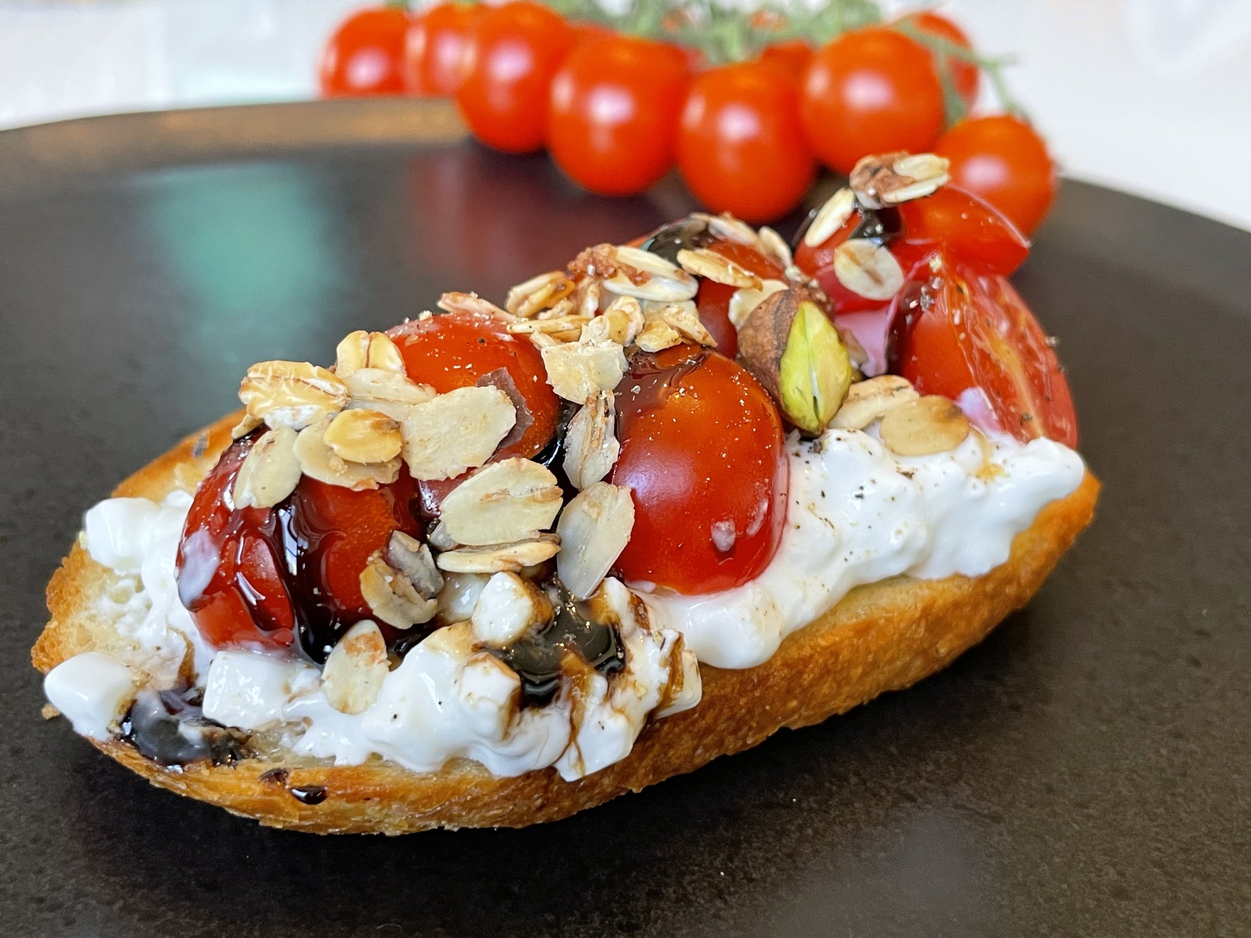 A slice of toast topped with creamy cheese, cherry tomatoes, chopped almonds, and drizzled with balsamic glaze, with a cluster of cherry tomatoes in the background.