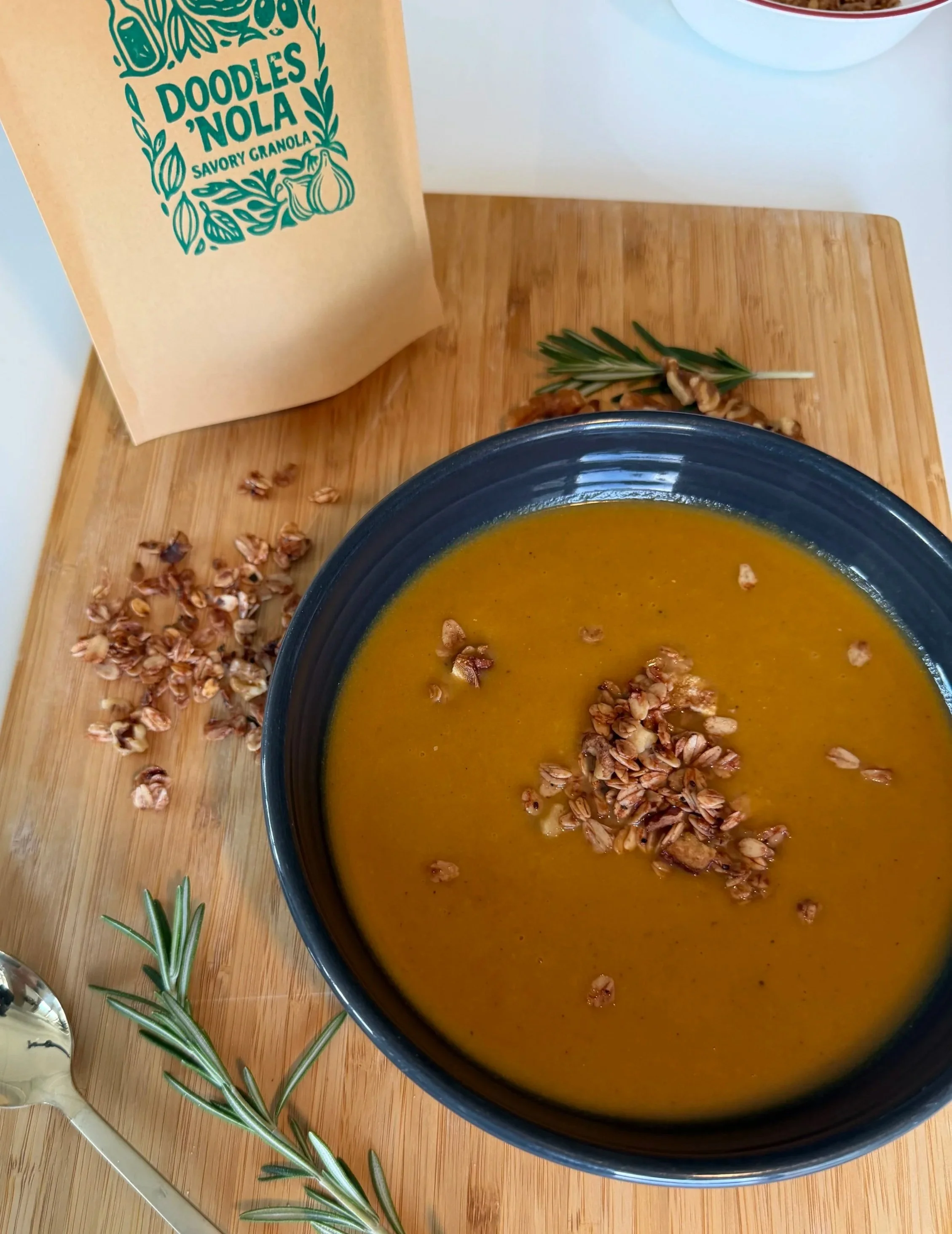 A bowl of orange soup topped with granola, placed on a wooden cutting board with sprigs of rosemary. In the background, a package labeled 'Doodles 'Nola savory granola' is visible on a white surface.