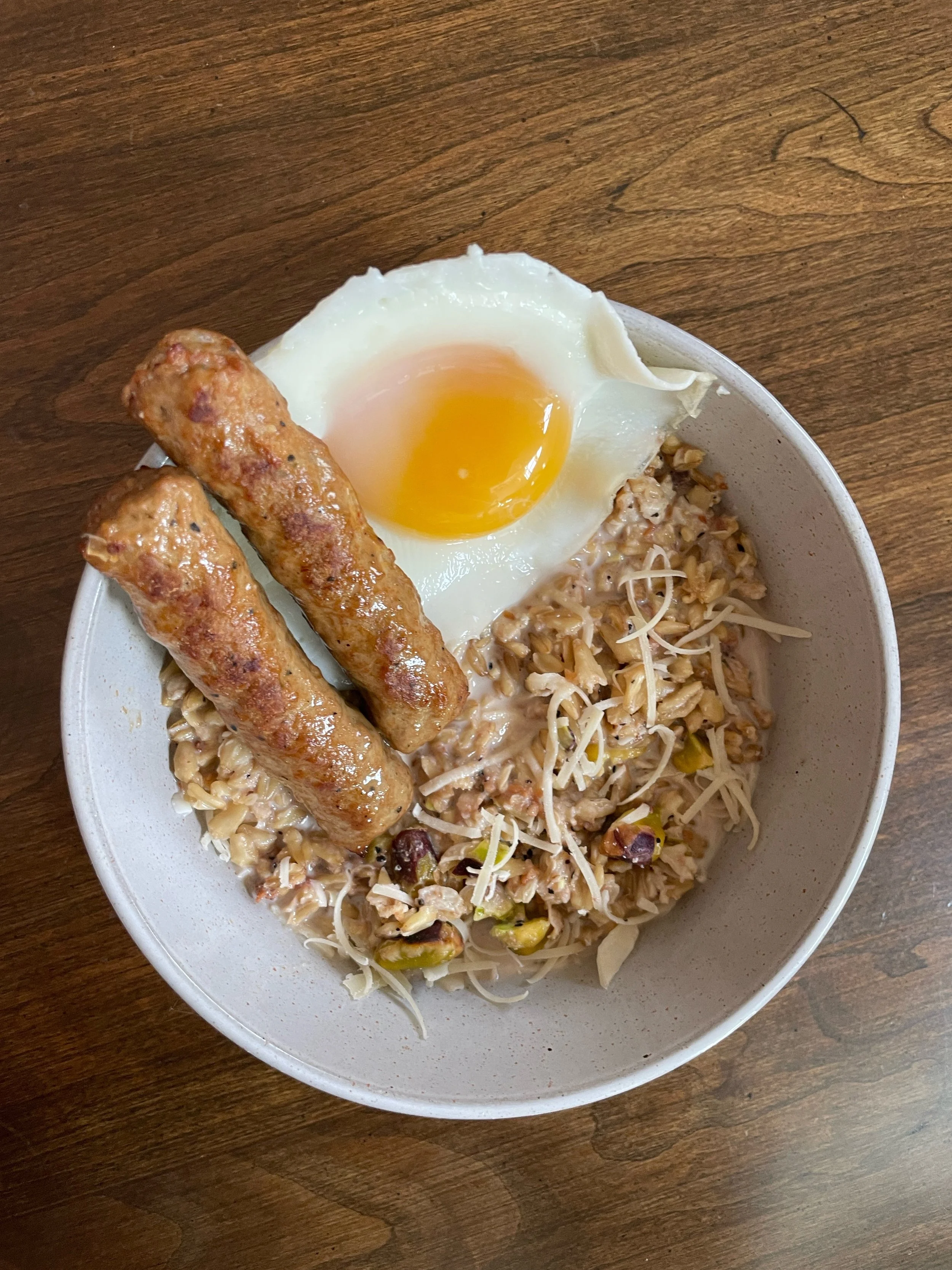 A bowl of rice or pasta with shredded cheese, topped with a fried egg, two sausage links, and a side of cooked vegetables.