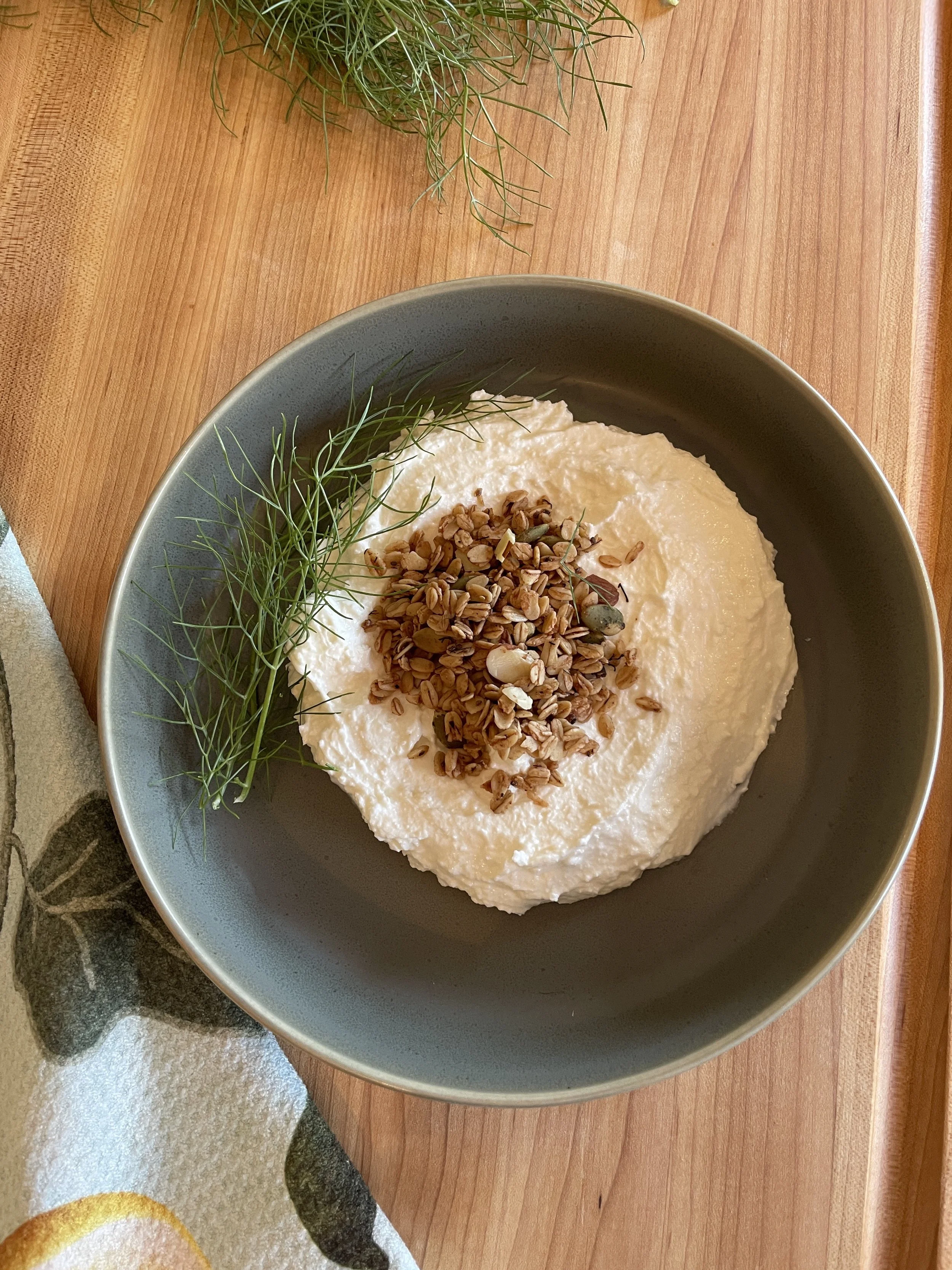 A bowl of creamy white hummus garnished with sunflower seeds and fresh dill, placed on a wooden surface with additional dill sprigs nearby.