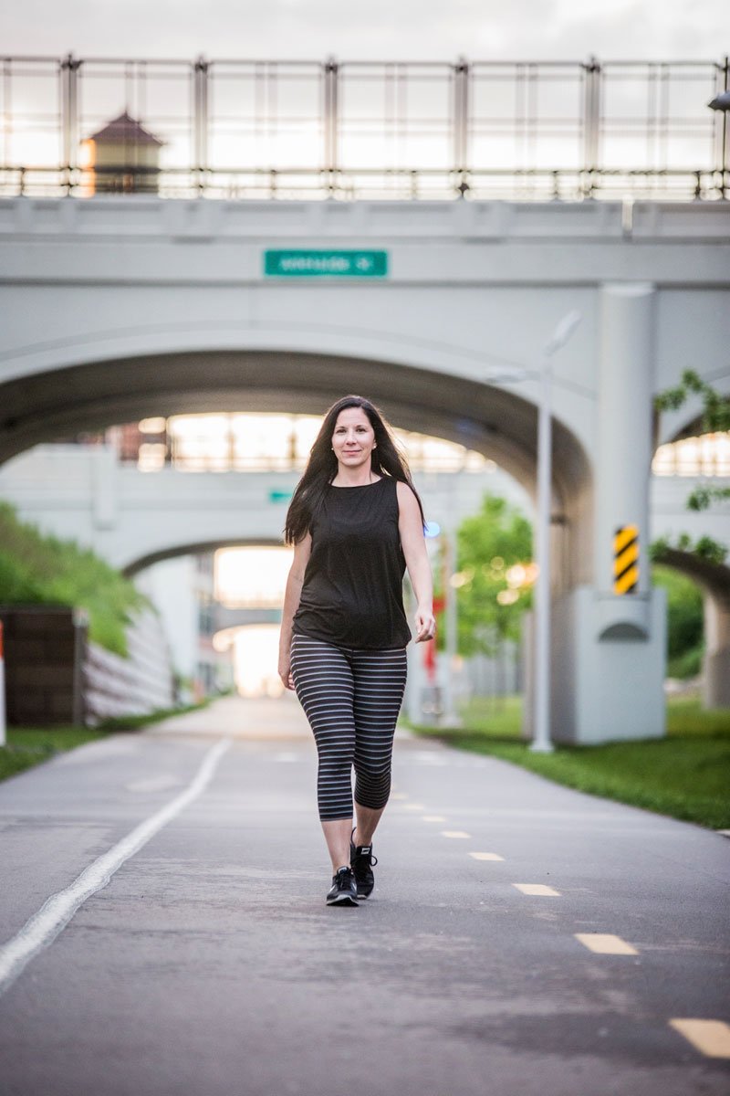 A woman, Melissa Boudreau, walking on a paved trail under a bridge, wearing a black sleeveless top and striped leggings, with greenery on the sides and a sunset sky in the background.
