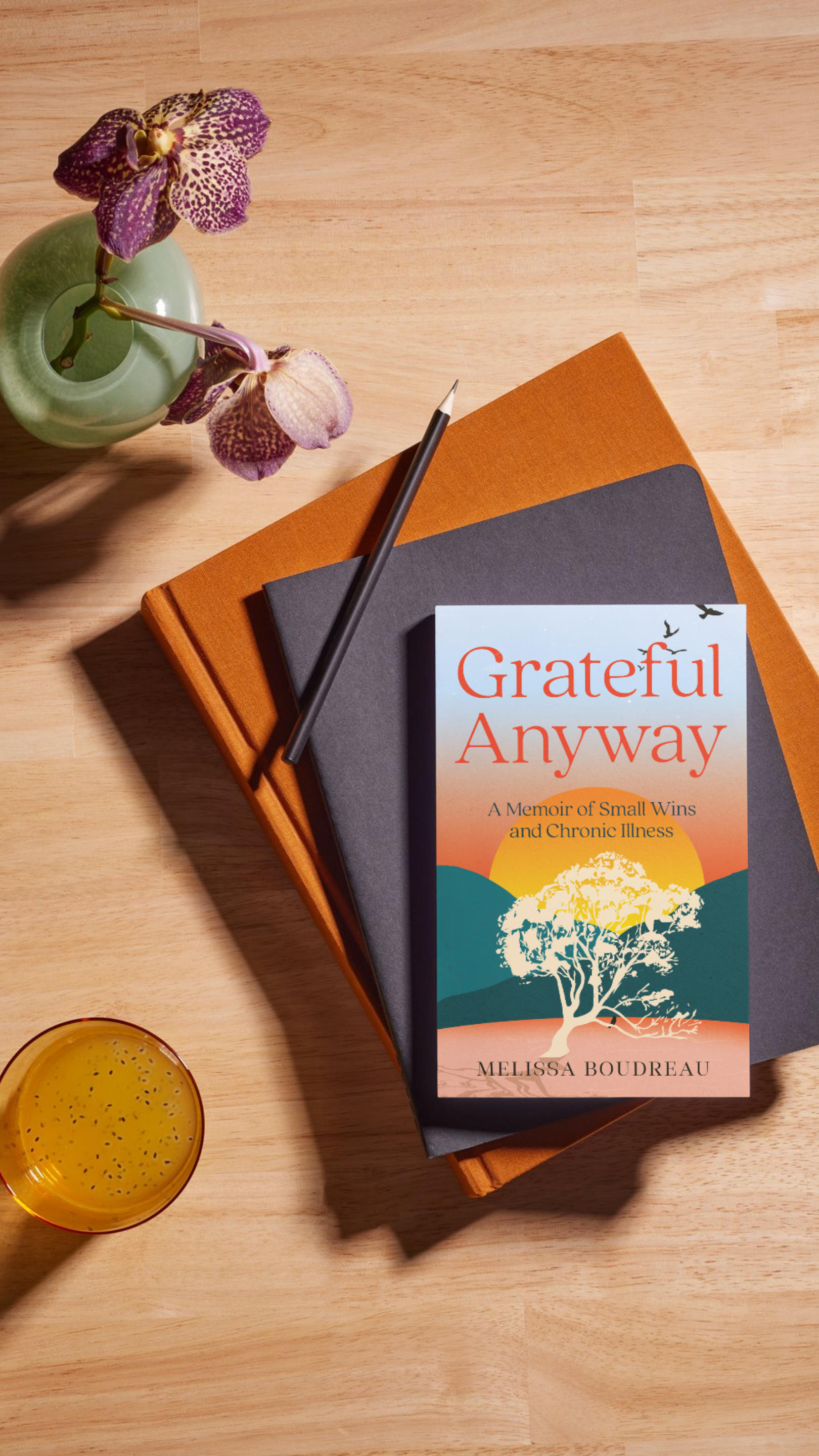 A book titled 'Grateful Anyway' by Melissa Boudreau resting on top of two closed notebooks, with a black pencil on top of the notebooks, a green vase with purple orchids, and a glass of yellow drink on a wooden surface.