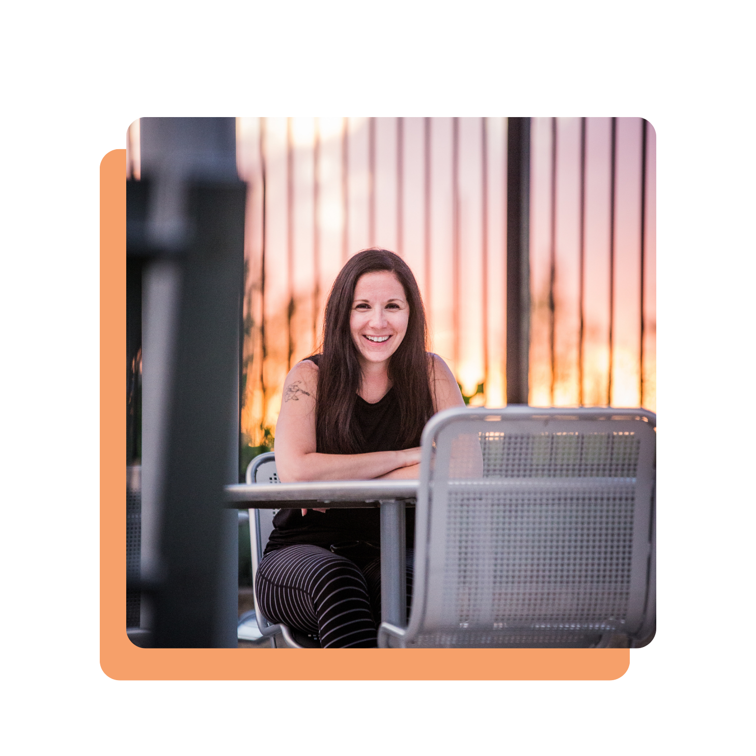A woman, Melissa Boudreau, with long dark hair sitting at an outdoor table, smiling at the camera during sunset, with a metal fence and sunset sky in the background.