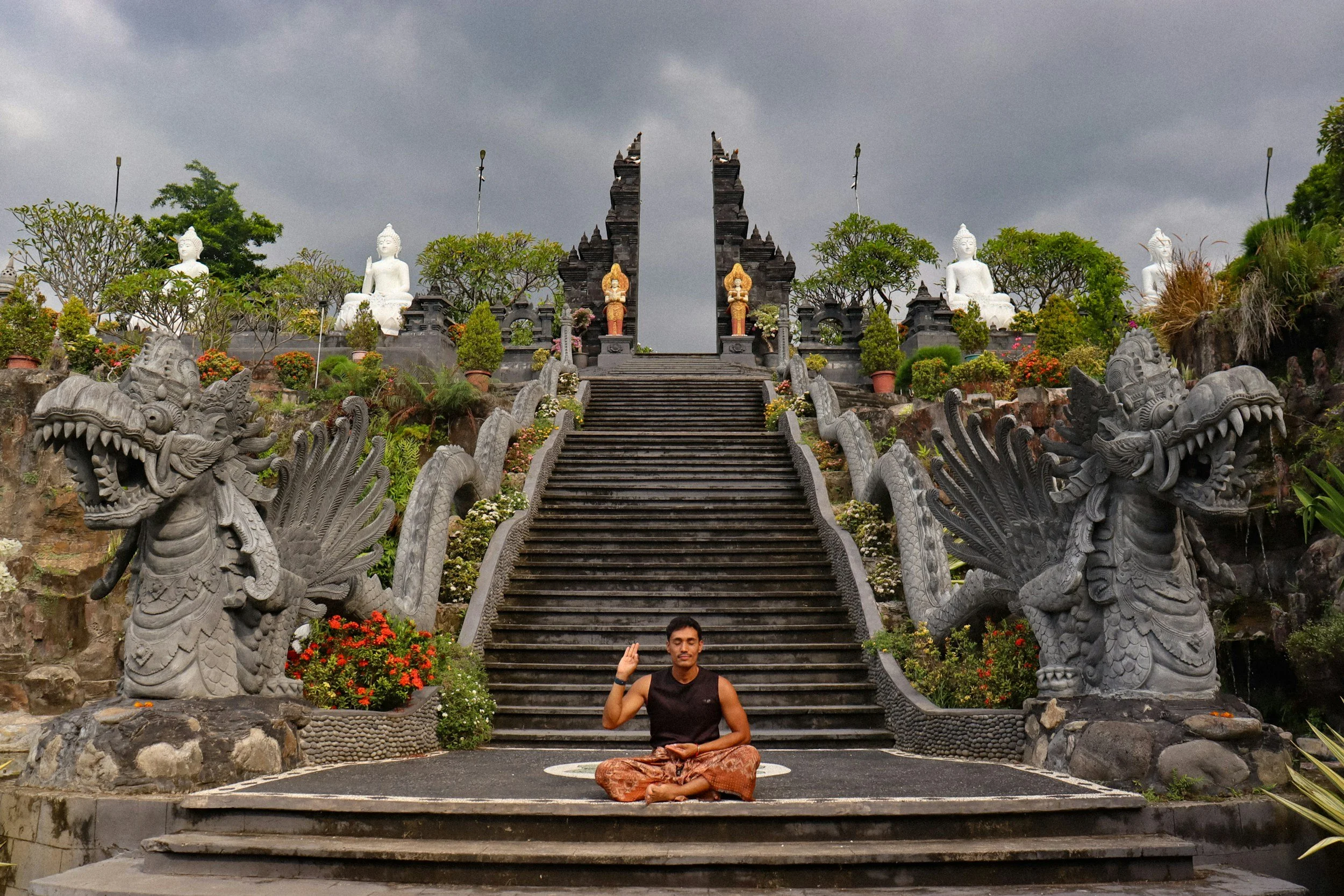 A person in a meditation pose sitting cross-legged at the bottom of stone staircase between carved stone dragon sculptures. The stairs lead up to a temple entrance with white Buddha statues on either side and two gold and red statues in the middle, surrounded by greenery and plants under a cloudy sky.