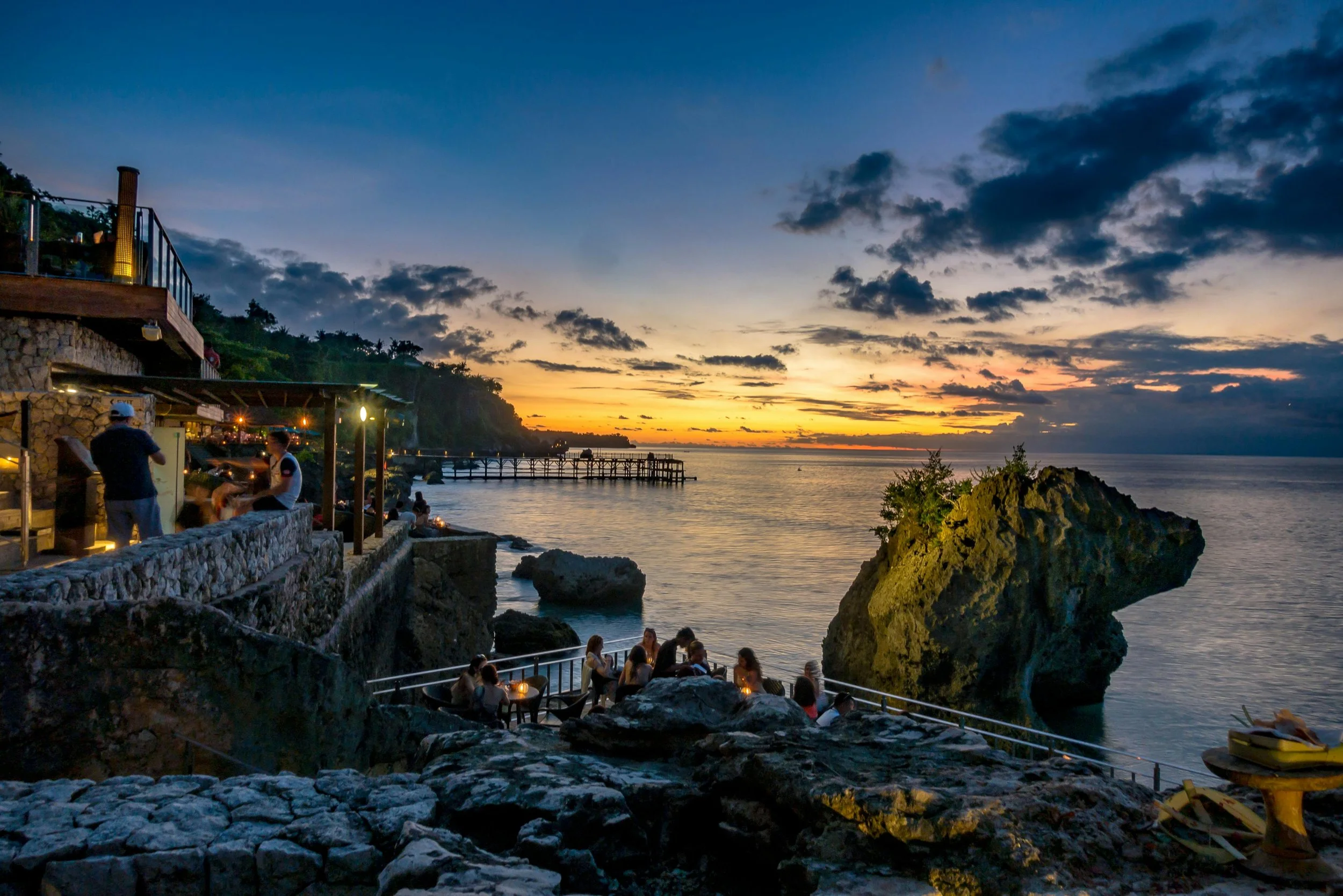 A seaside restaurant at sunset with people dining on a stone terrace overlooking the ocean, cloudy sky, and distant pier.