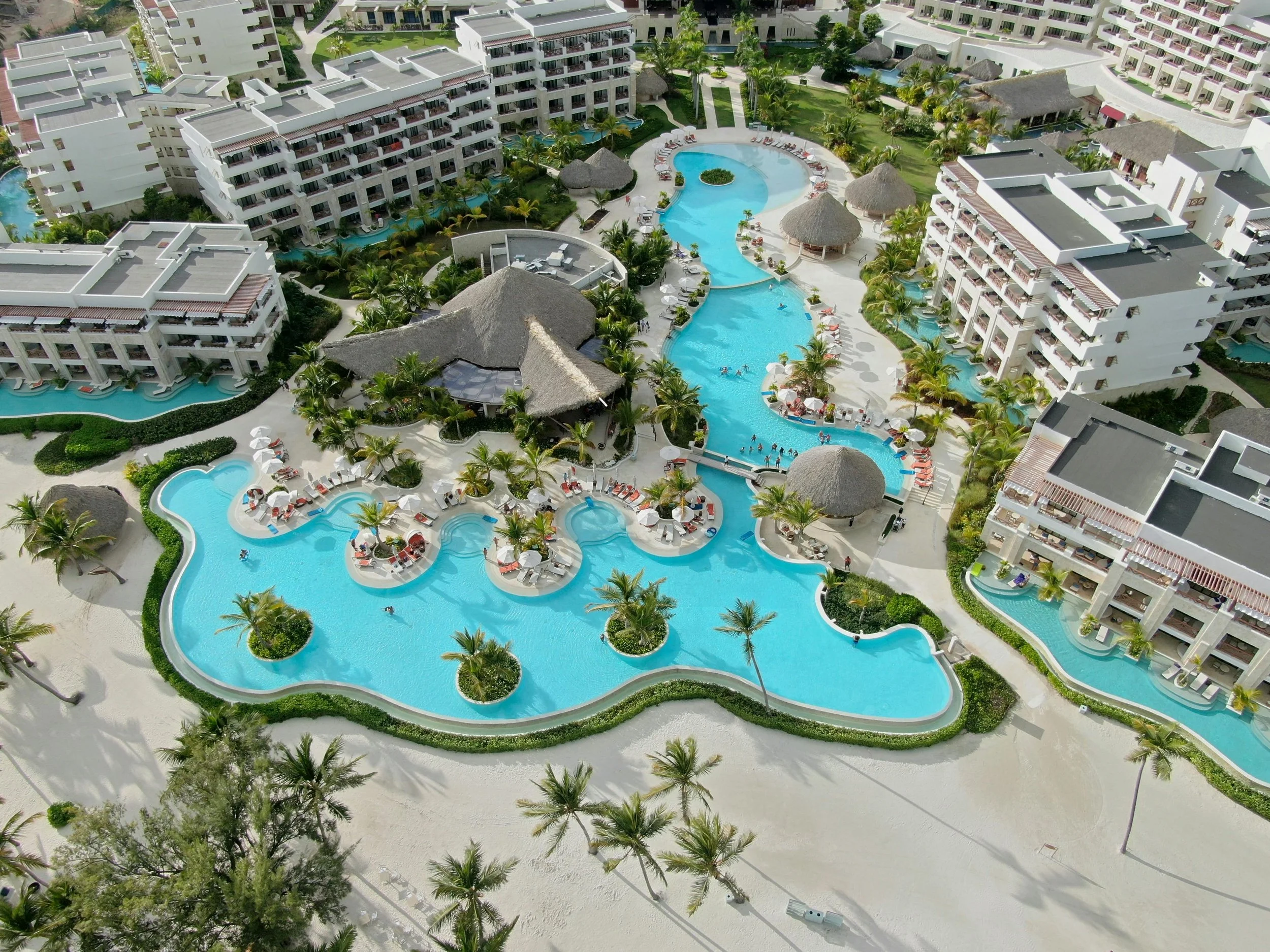 Aerial view of a resort with multiple white buildings surrounding a large curvy swimming pool with poolside lounge chairs and umbrellas, tropical palm trees, thatched-roof cabanas, and green landscaped areas.