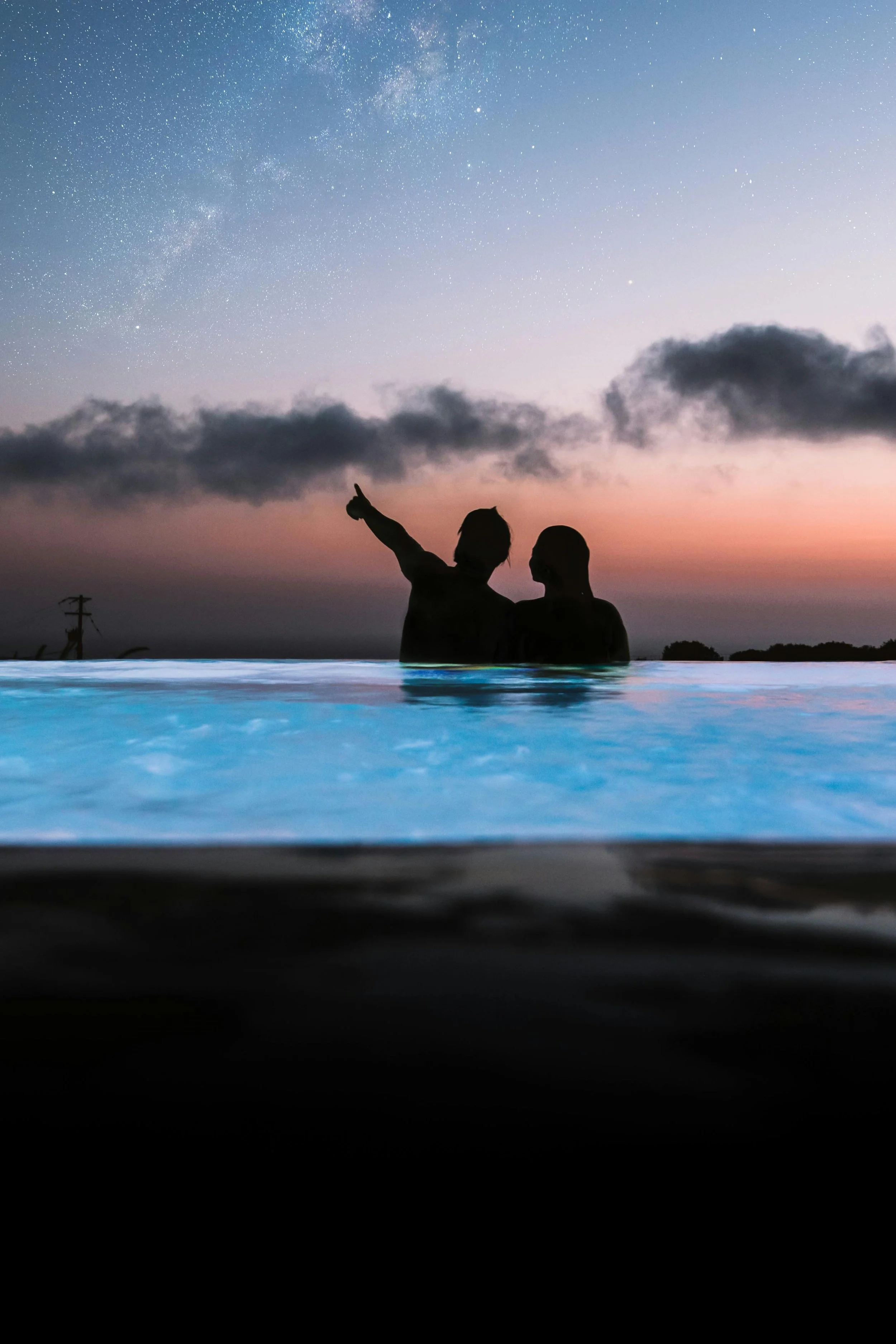 Two people silhouette sitting in a swimming pool during sunset, pointing at the starry night sky with clouds and the Milky Way.
