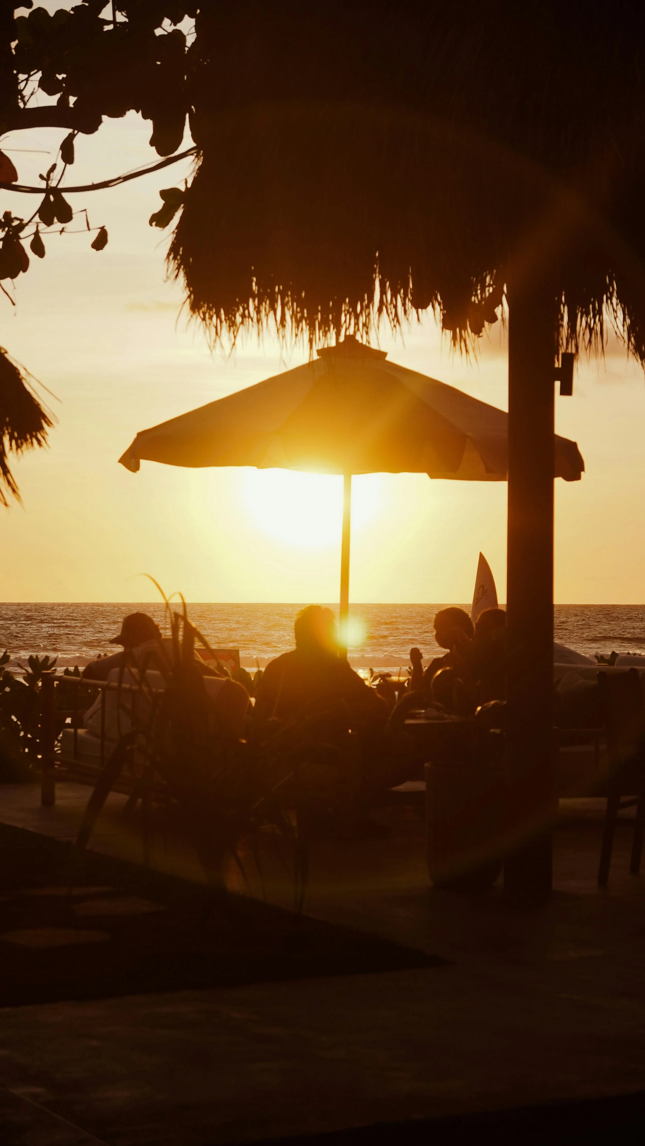 People sitting at a beachside café during sunset with a straw-roofed hut, an umbrella, and the ocean in the background.