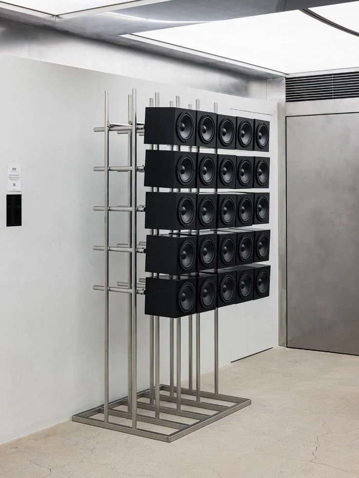 A modern speaker display with multiple black speakers mounted on a metal frame against a white wall.