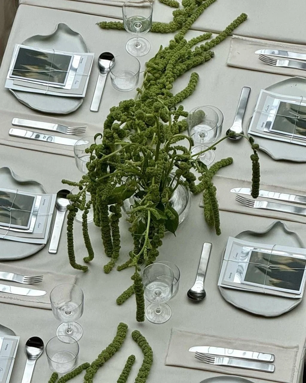 A table setting with plates, cutlery, wine glasses, and a centerpiece made of green, elongated, knobby plant stems arranged in a glass vase.