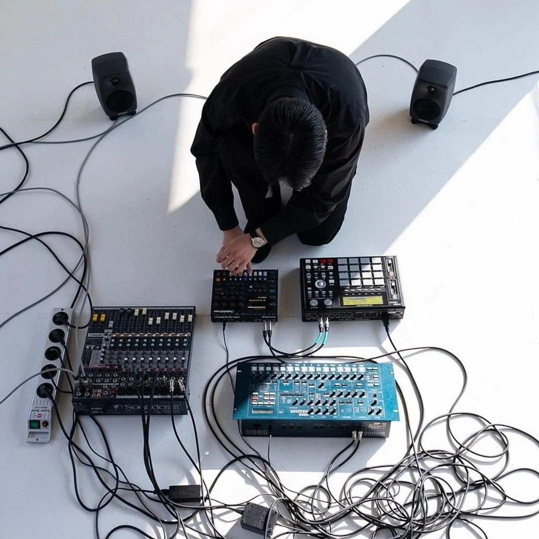 A person dressed in black sitting on the floor surrounded by electronic music equipment, including a mixer, drum pad, synthesizer, and speakers, with tangled cables around.