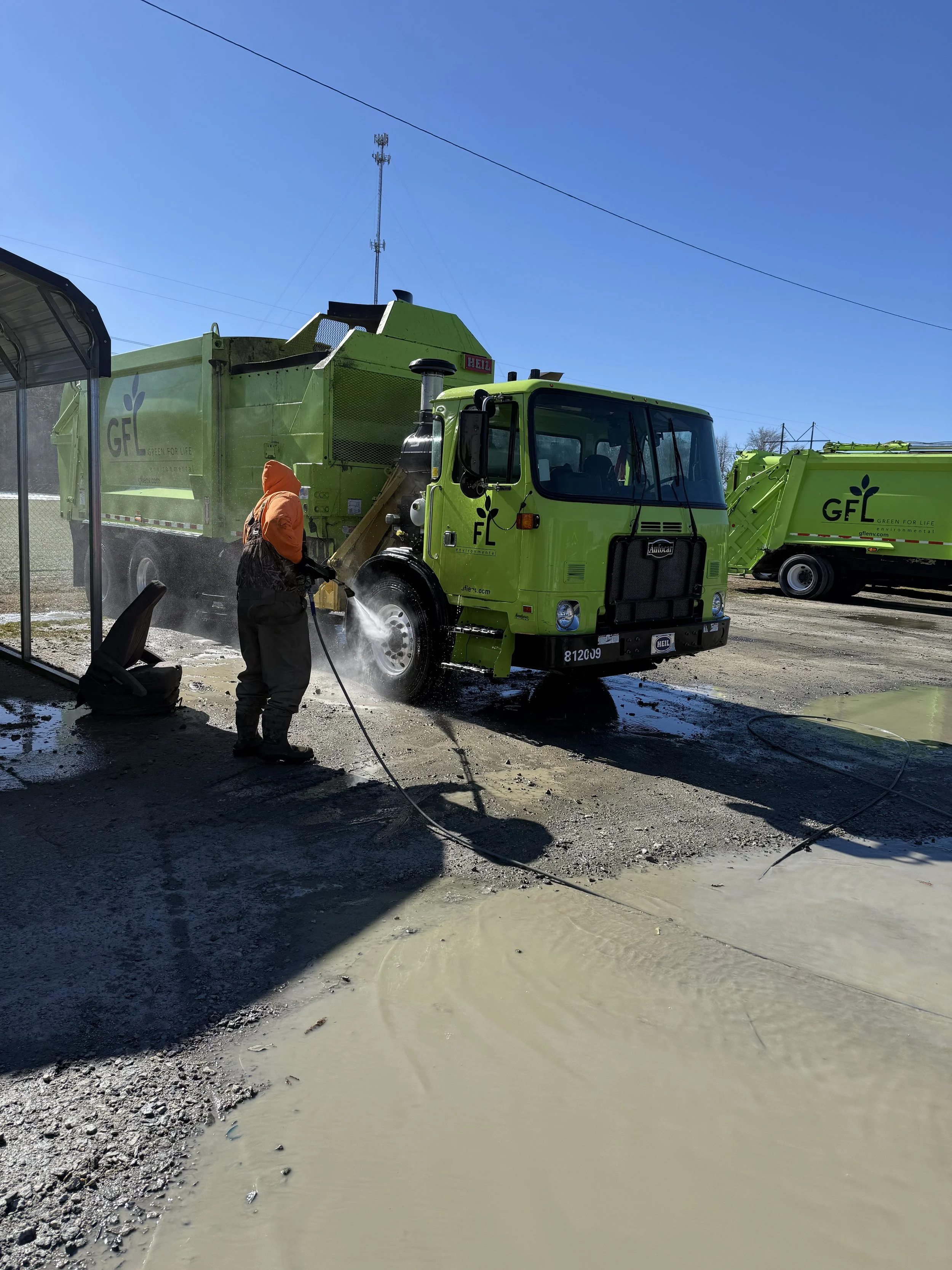 Worker wearing orange hoodie and black coveralls pressure washing a bright green street sweeper vehicle outdoors on a sunny day.