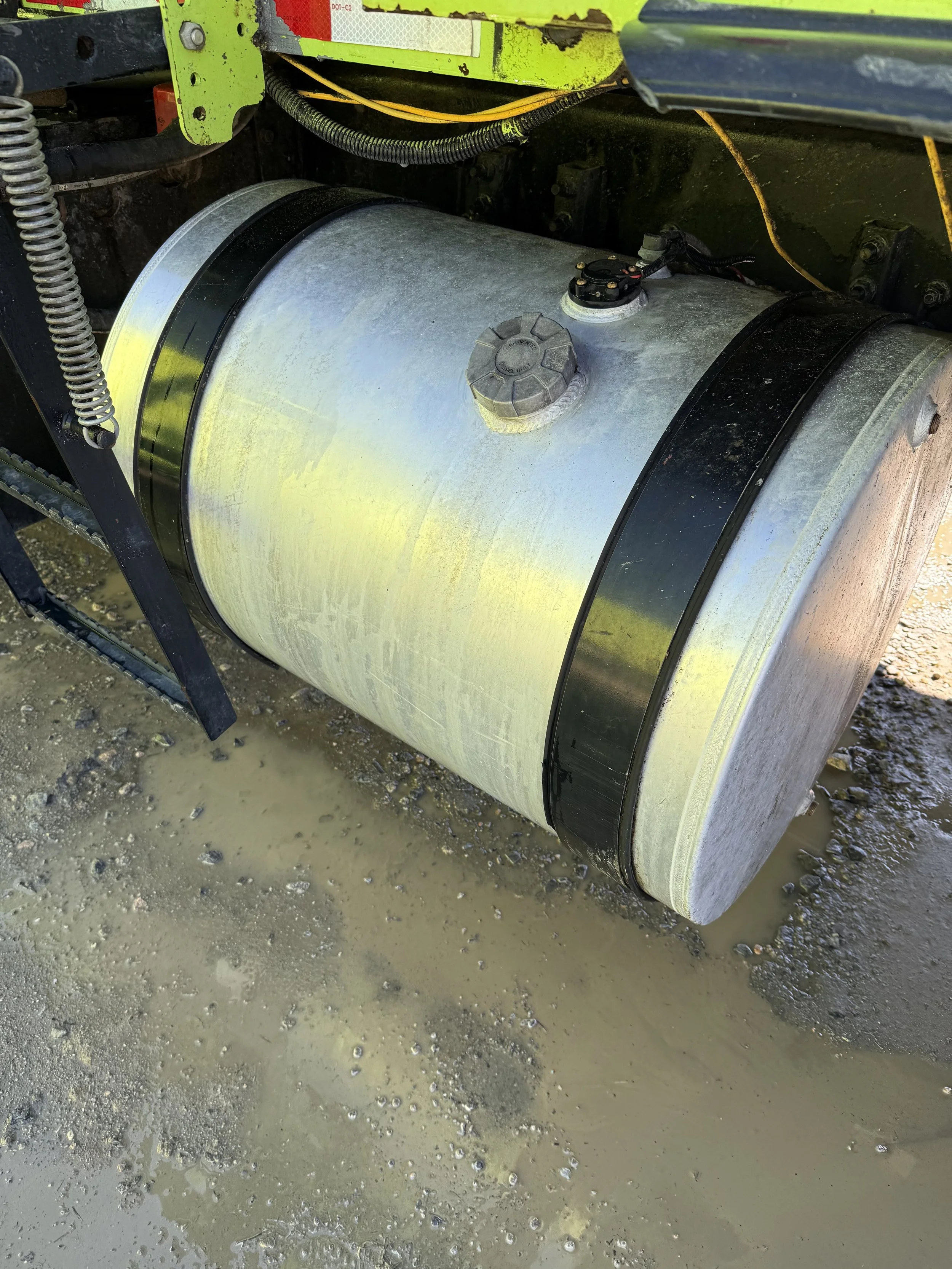 Close-up of a large metallic cylindrical tank on a muddy ground, with a black band around it, a cap on top, and electrical wires connected to its surface.