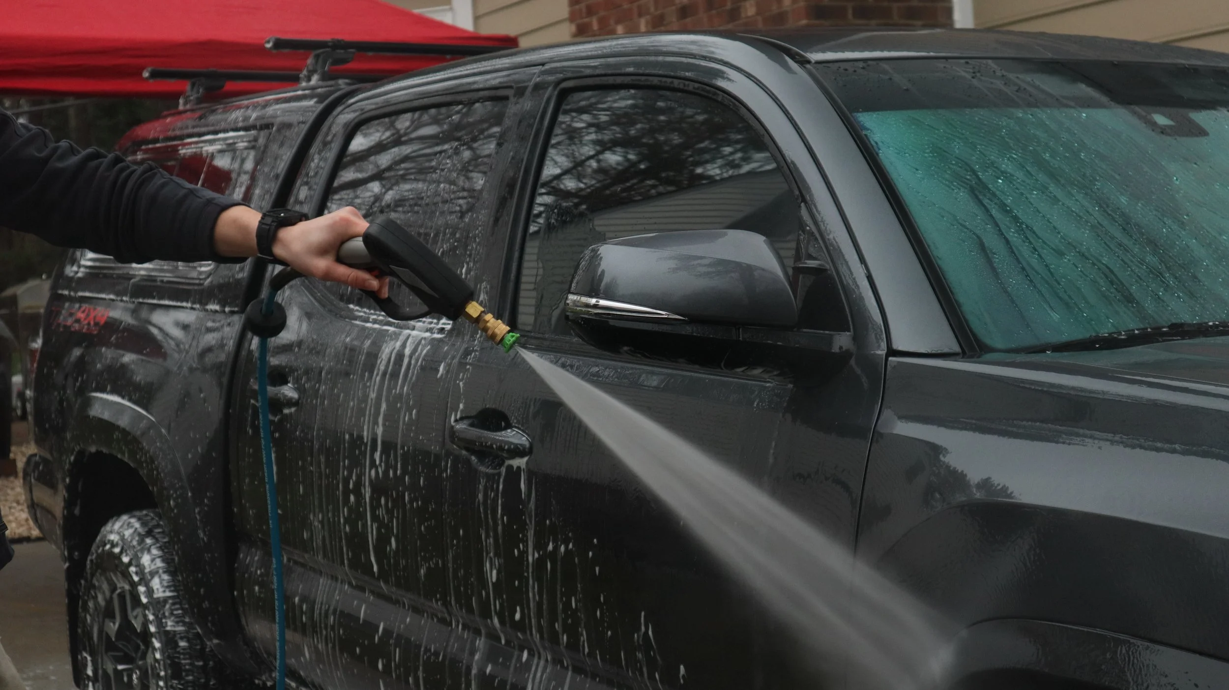 Person washing a black pickup truck with a pressure washer, soap and water on vehicle surface, windshield wipers, and side mirror visible, outdoors.