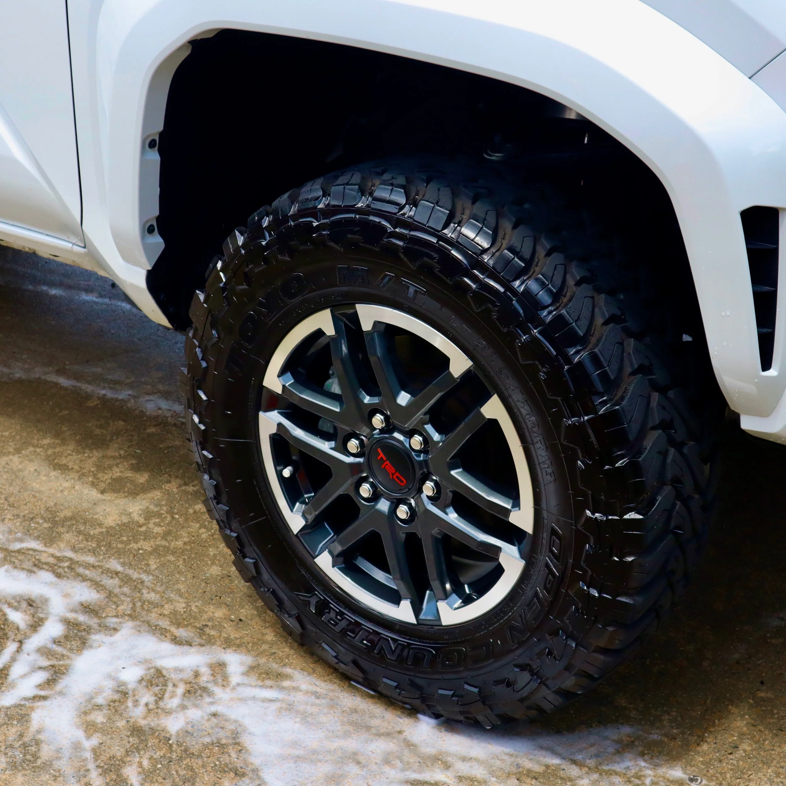 Close-up of a white vehicle wheel with large off-road tires, parked on a muddy surface with patches of snow.