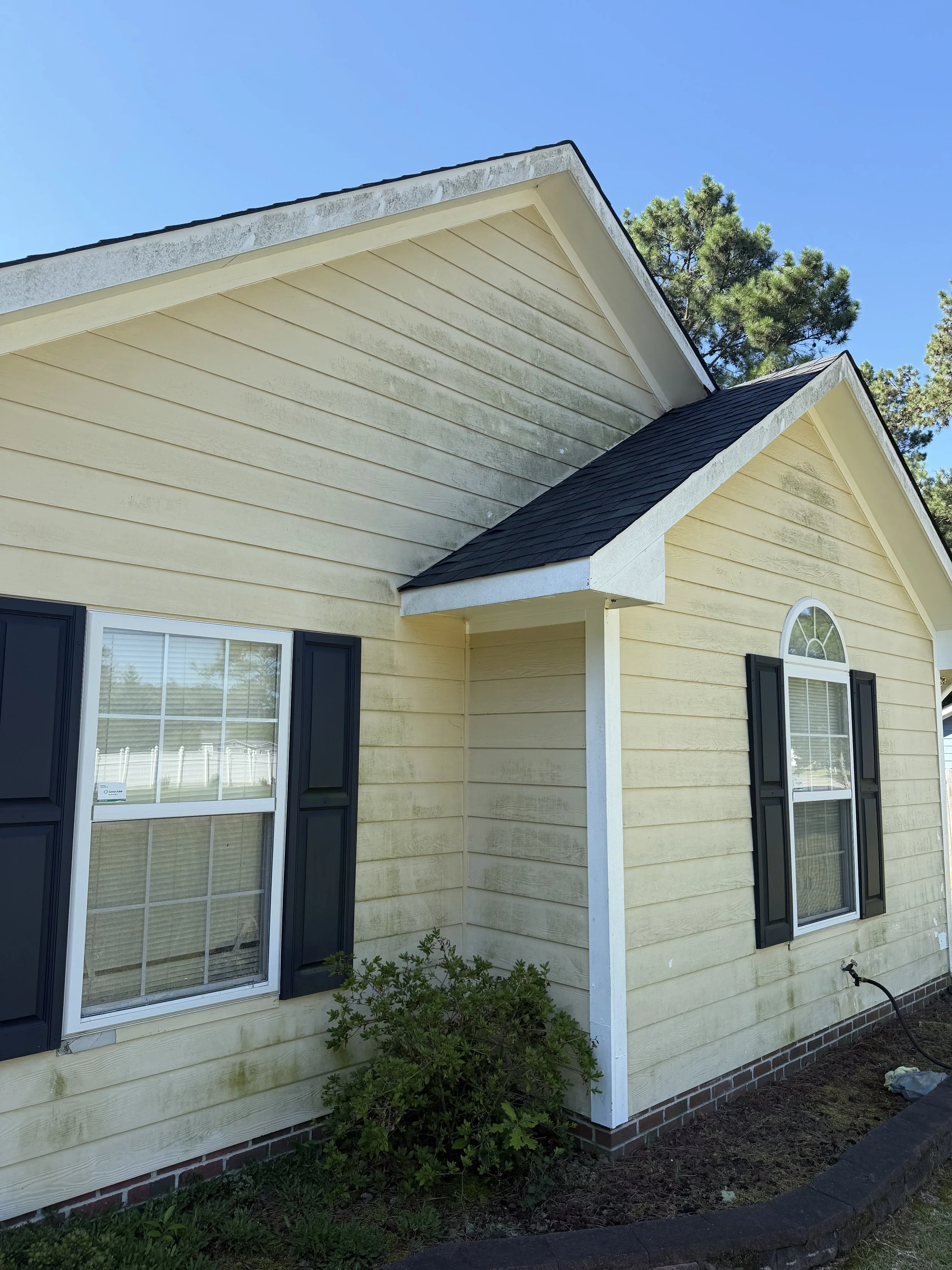 Exterior of a house with yellow siding, black window shutters, and a brick foundation, showing two windows with blinds and a part of a garden bed.