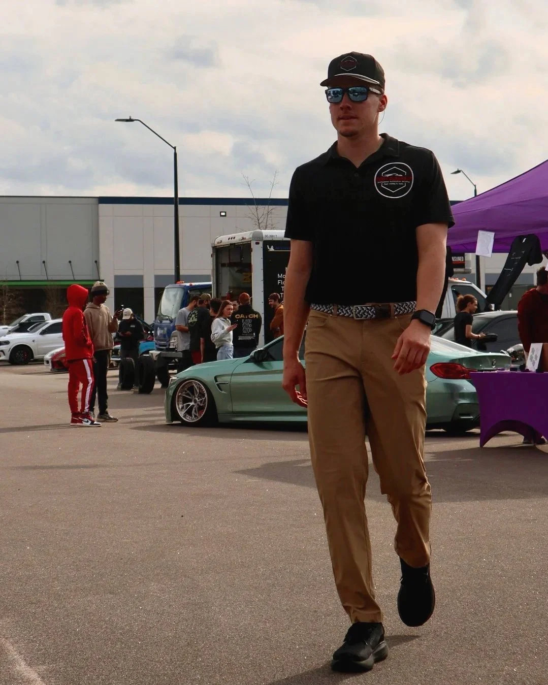 A young man walking at an outdoor car show wearing a black baseball cap, sunglasses, a black polo shirt, khaki pants, and black shoes. In the background, there are several people, a green sports car, and various tents and vehicles.
