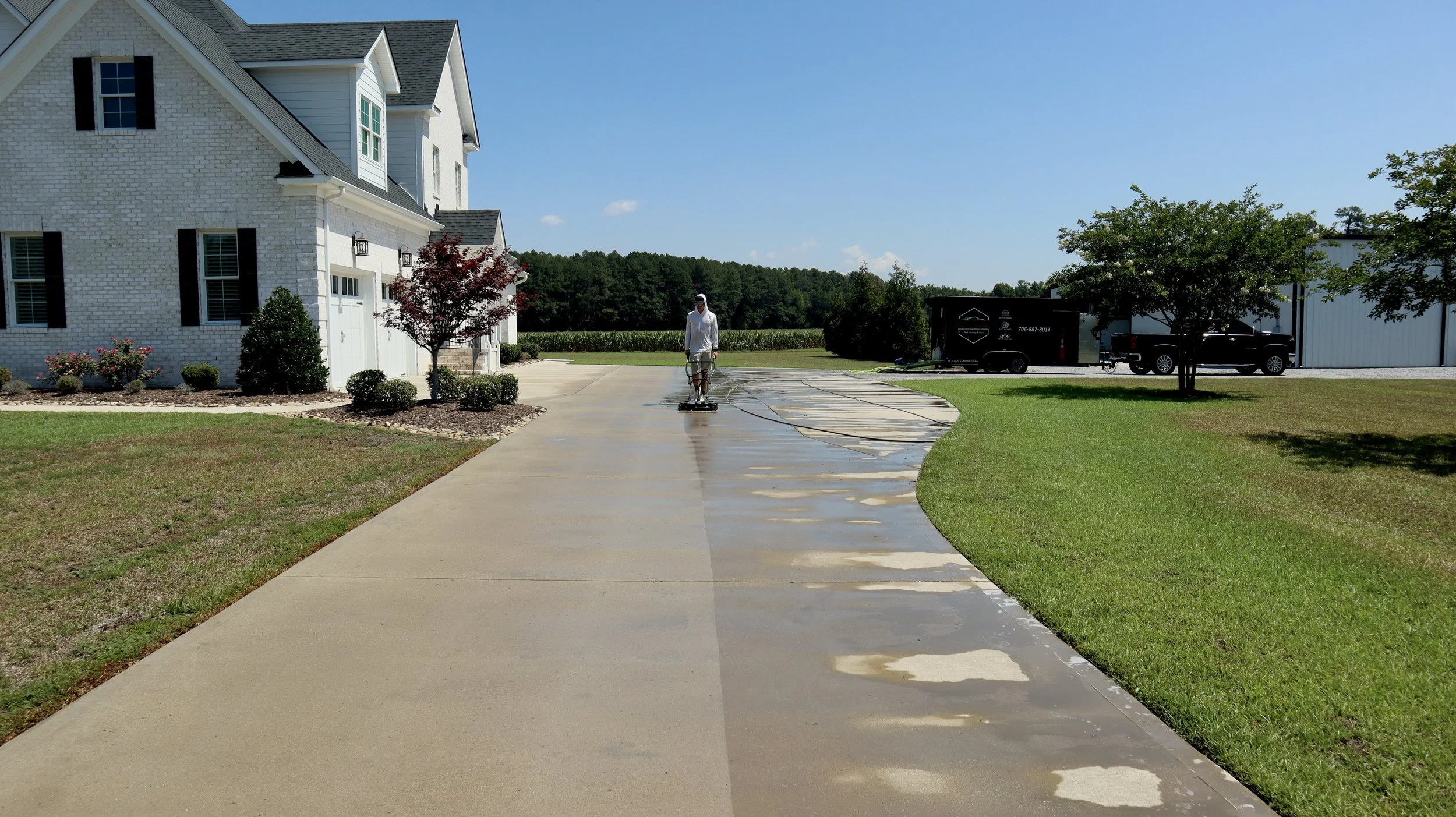 Person pressure washing a concrete driveway in front of a white house with landscaping and parked trucks in the background.