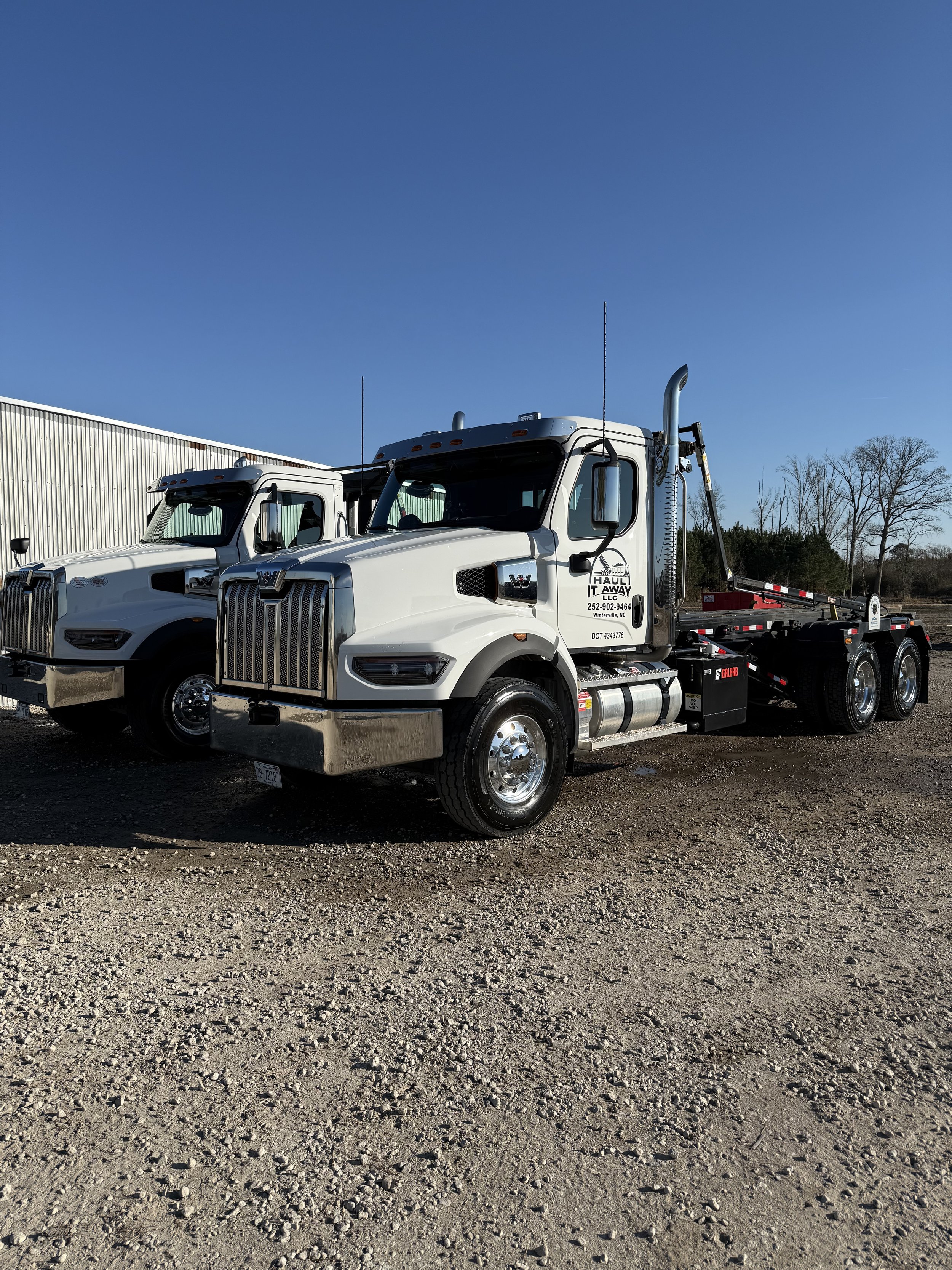 White flatbed tow truck with company signage, parked outdoors on gravel, with another similar truck, a building, and leafless trees in the background, under a clear blue sky.