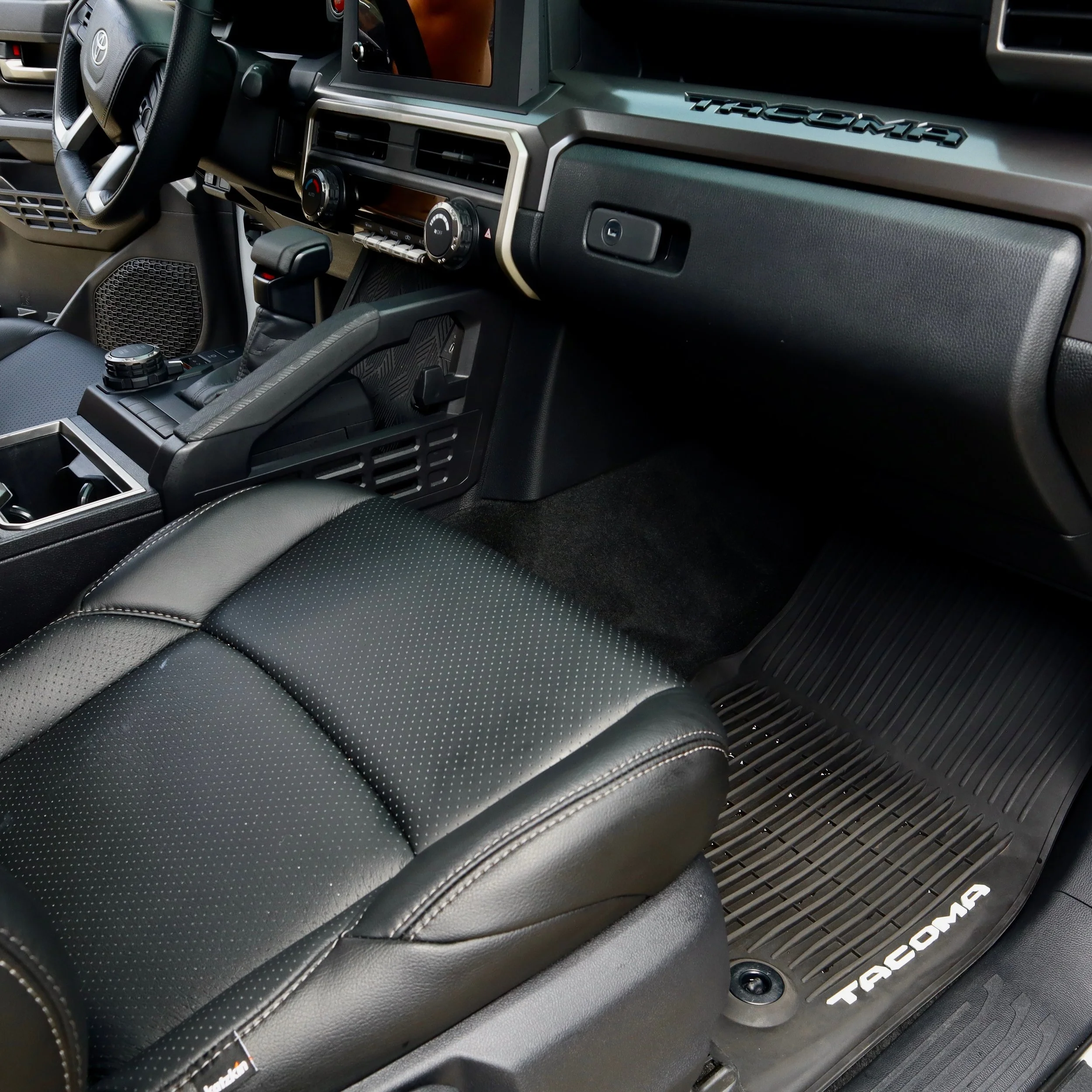 Close-up of the interior dashboard of a Toyota Tacoma truck, showing the steering wheel, air vents, control knobs, and part of the black leather seat.