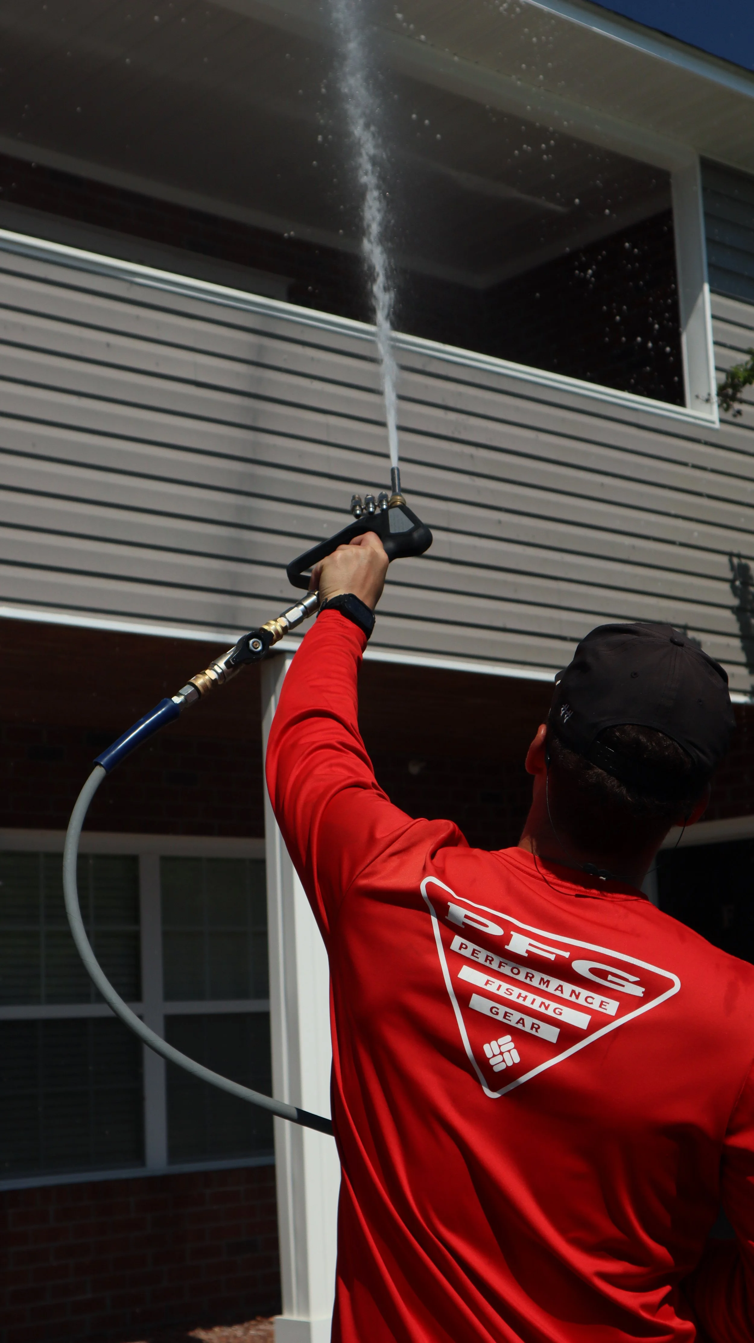 A person wearing a red shirt with a logo, holding a spray nozzle, cleaning the exterior of a house's window frame from below.