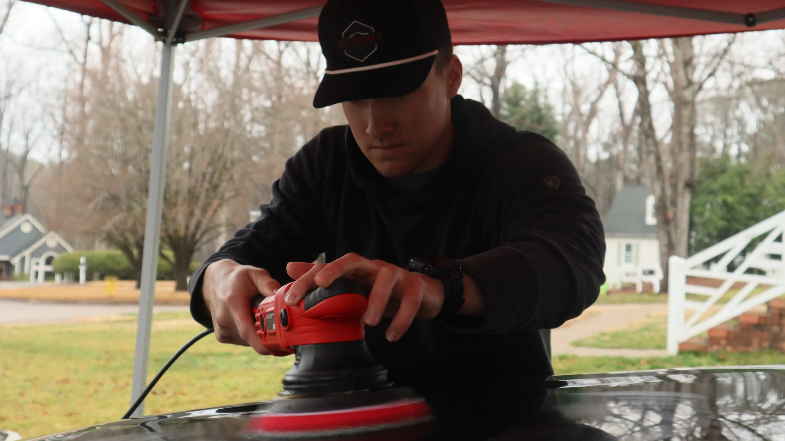A person in a black hoodie and cap polishing a black car with a red electric buffer under a canopy, with suburban houses and trees in the background.