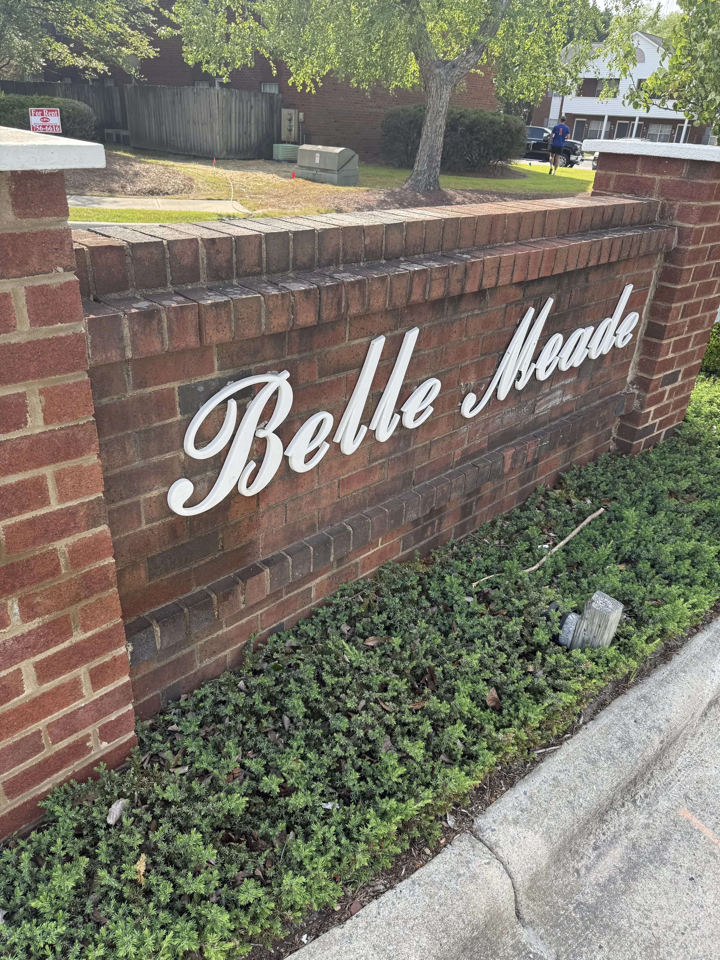 A large brick sign with white cursive lettering reading 'The Reserve' is surrounded by low green shrubs along a sidewalk. Shadows from trees are seen overhead.