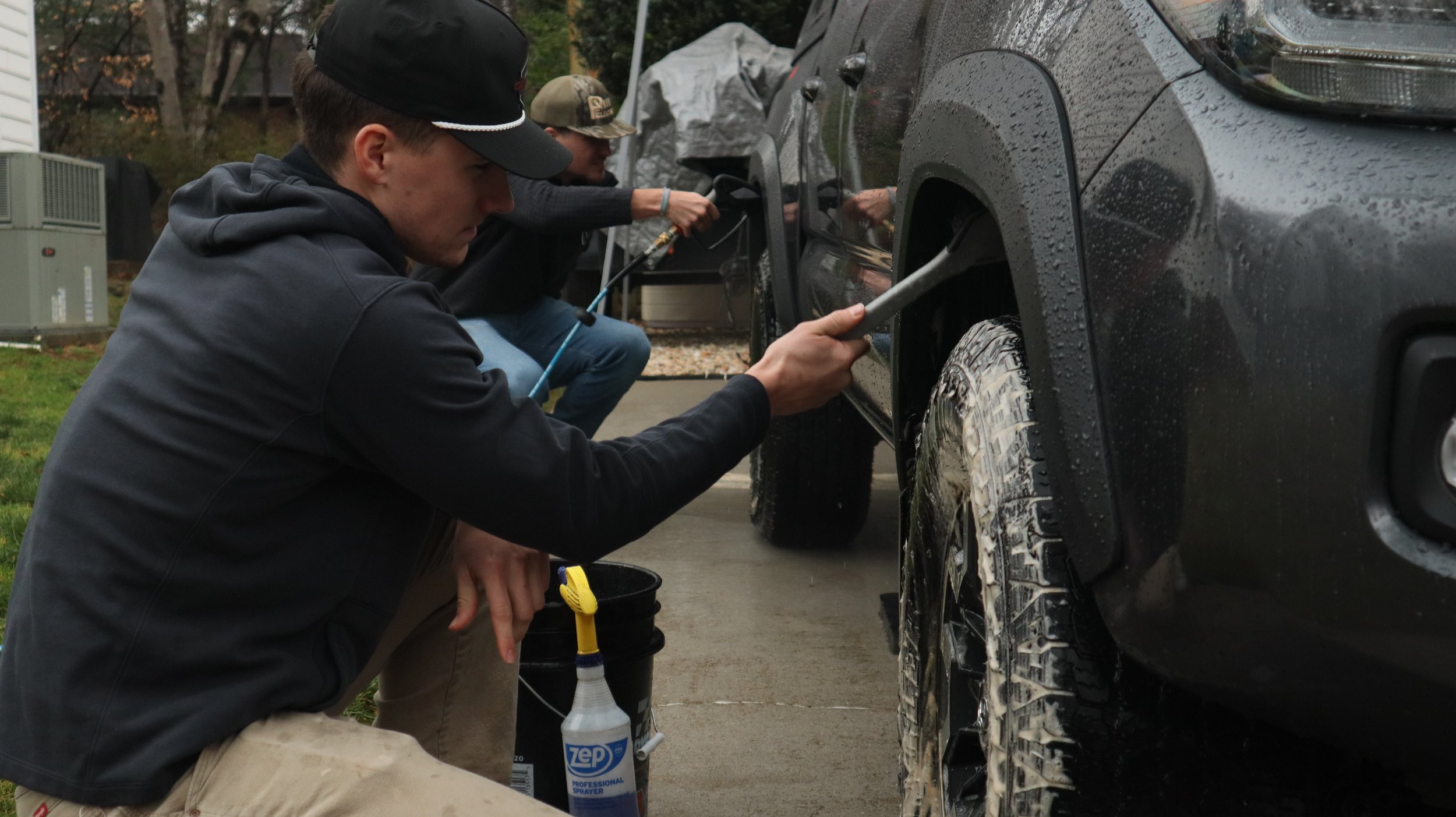 Three young men washing a black vehicle outdoors on a rainy day, with trees in the background. They are scrubbing the car's tires and body, using cleaning supplies and equipment.