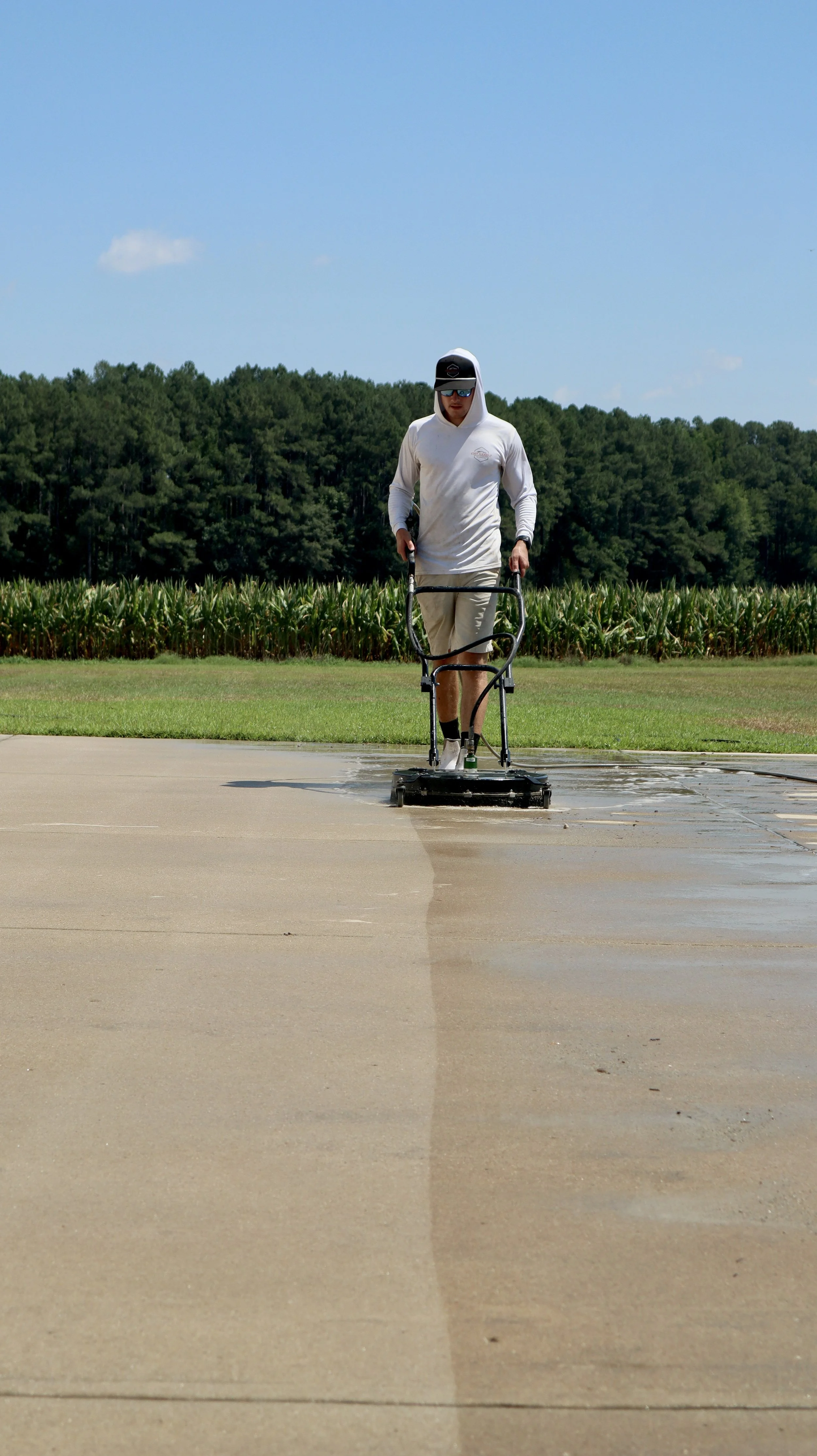 Man wearing sunglasses, hoodie, and shorts using a pressure washer to clean a concrete sidewalk outdoors, with a field and trees in the background.