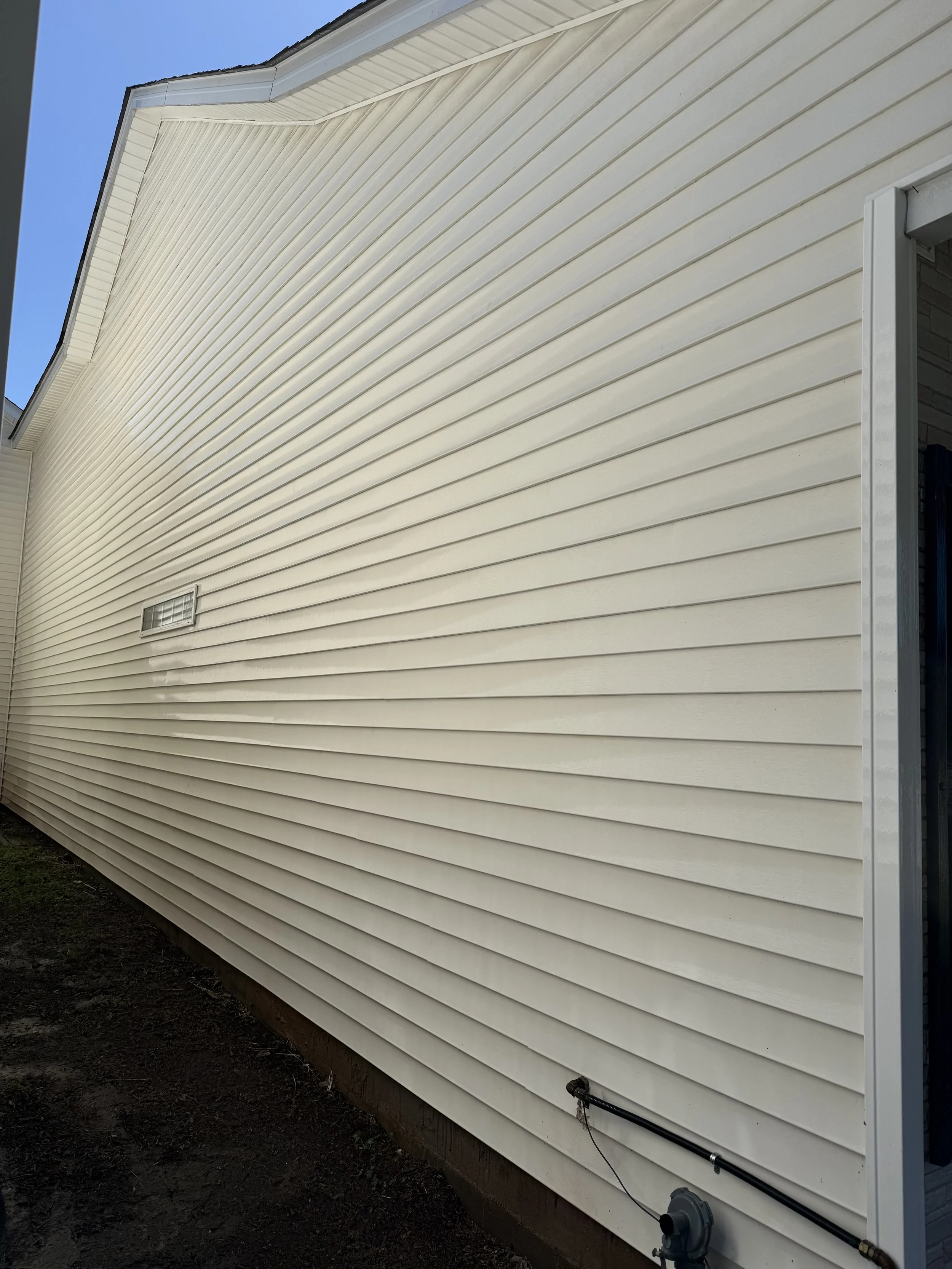 Siding of a house with beige horizontal vinyl panels and a small vent, located next to a sidewalk or garden bed.