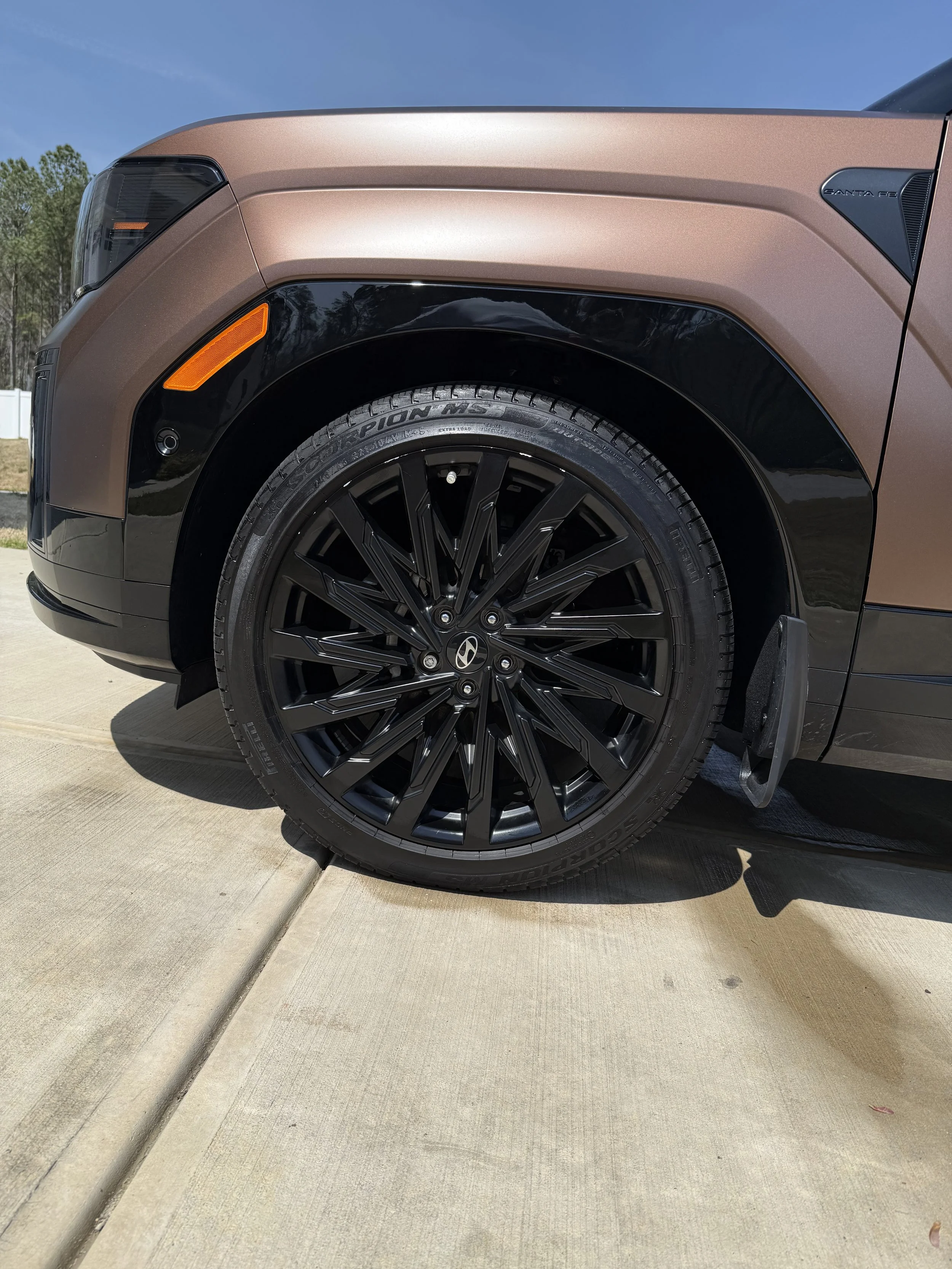 Close-up of the front left side of a brown and black Hyundai Santa Fe SUV, showing the wheel, tire, and part of the car body.