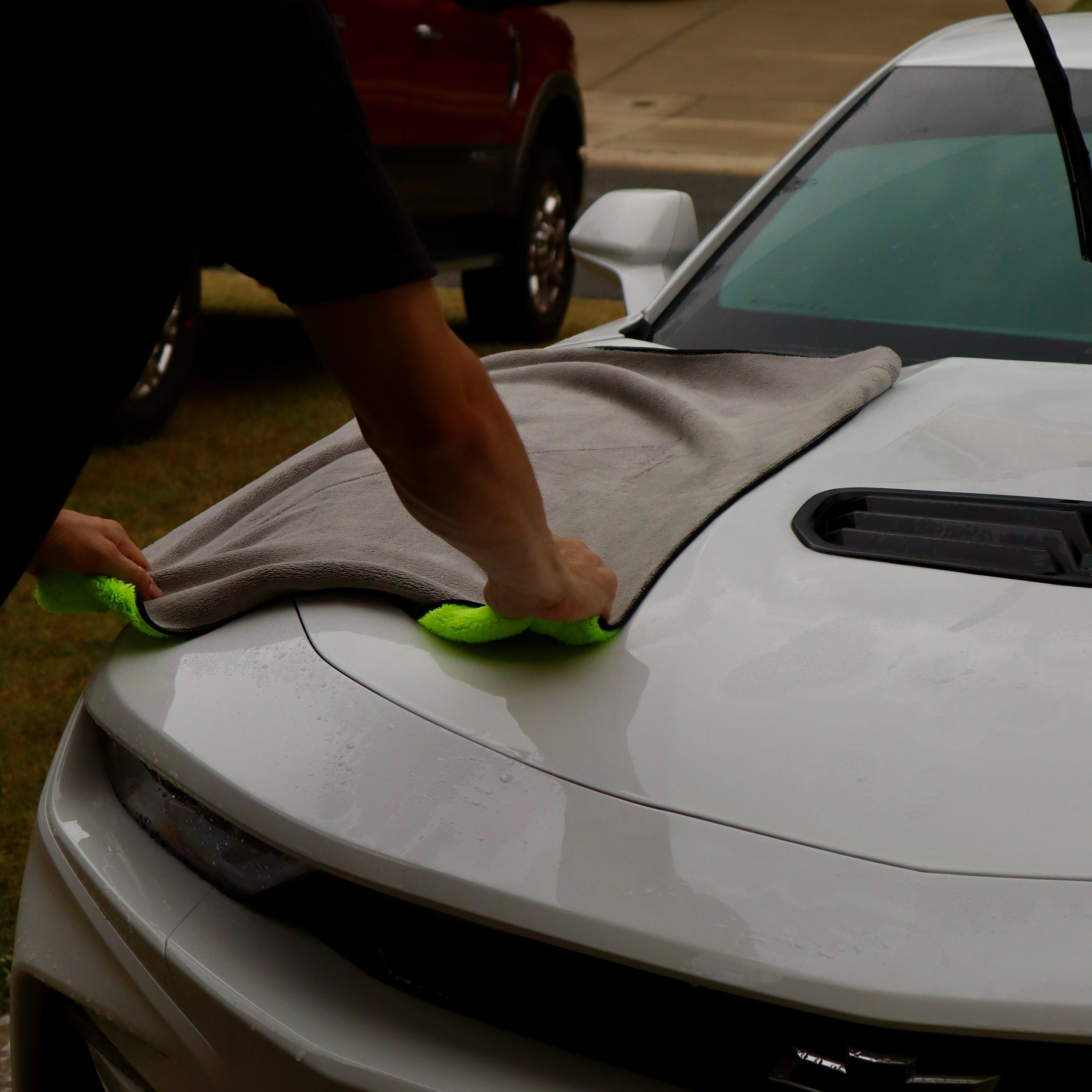 Close-up of the front end of a white sports car, showing part of the hood, windshield, and side mirror, parked outdoors with a blurred background.