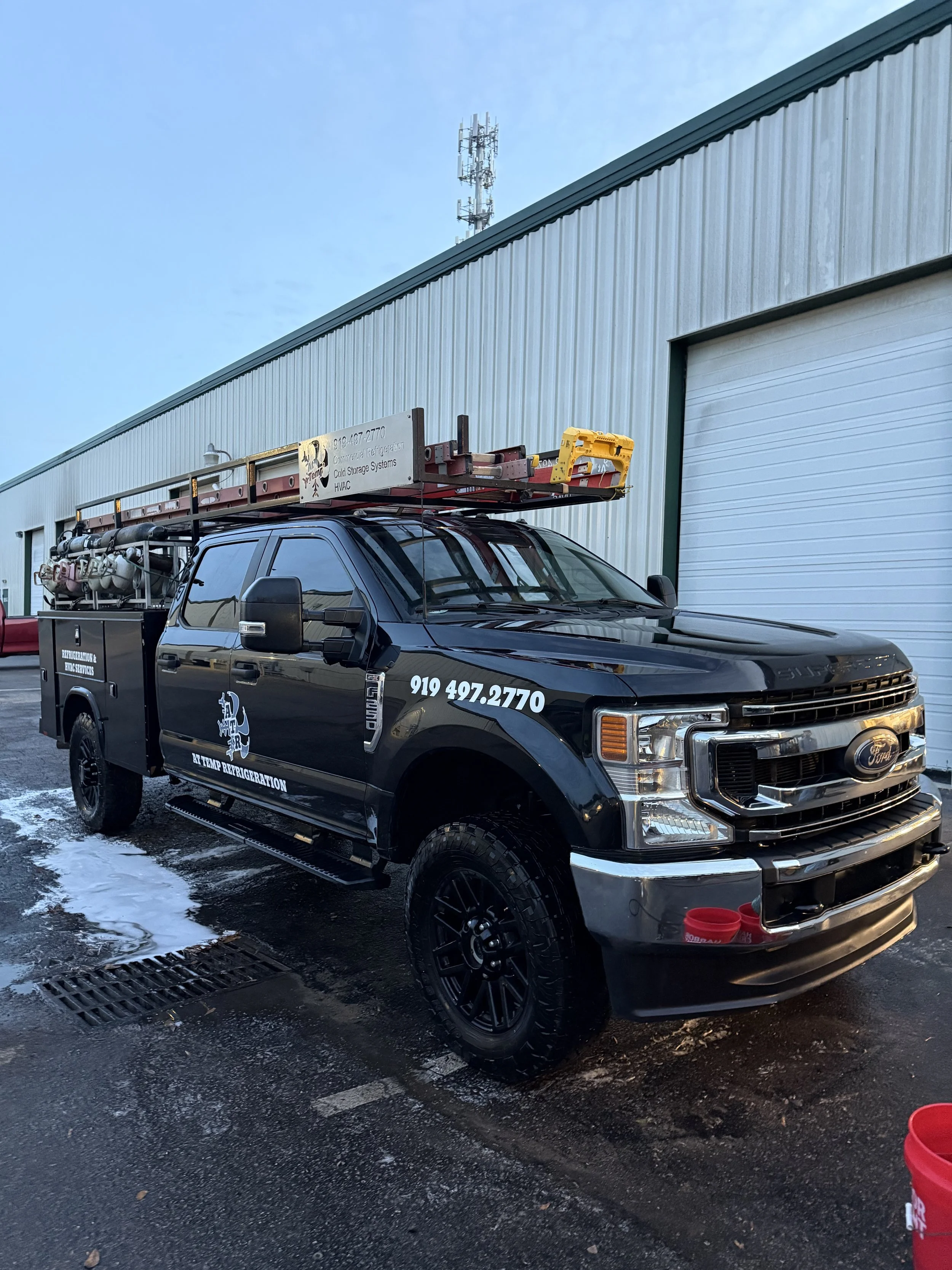 Black service truck with ladder and equipment on top parked outside a light blue industrial building.