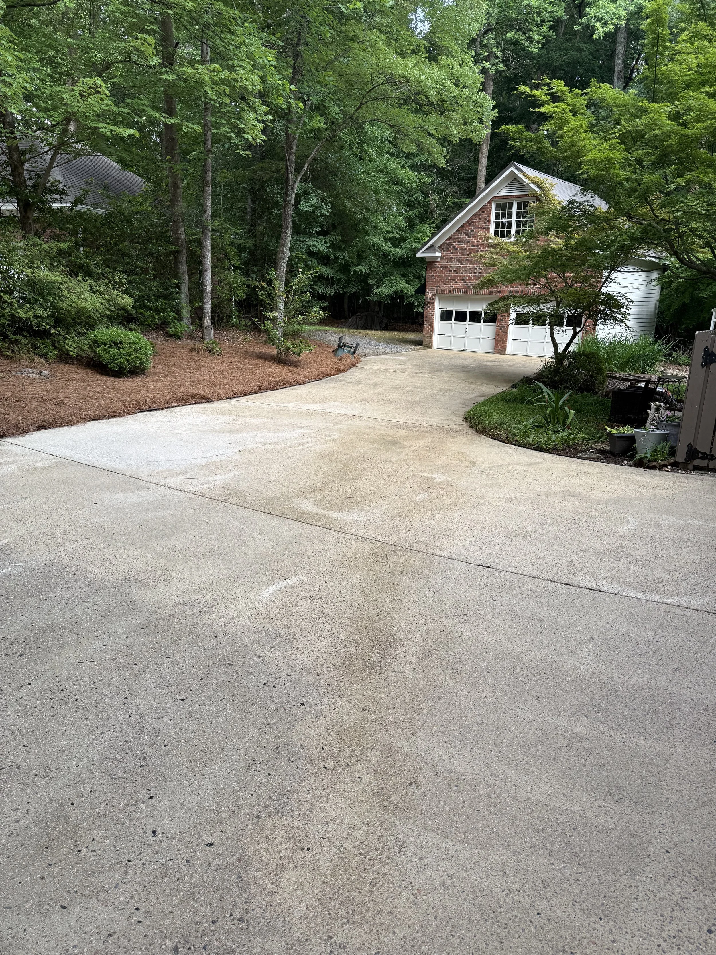 A curved concrete driveway leading to a two-car garage attached to a brick and siding house, surrounded by trees and landscaping.